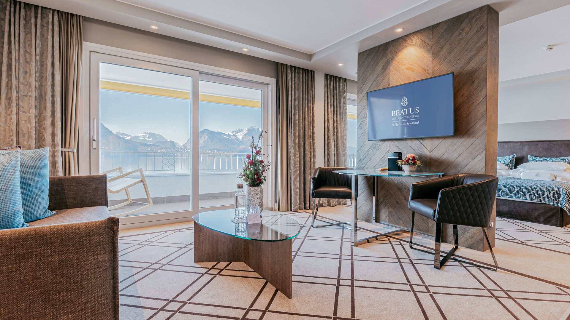Eiger Junior Suite at the Hotel BEATUS Room with carpet, glass table, view of the balcony and mountains, in front of a partition wall a small table with comfortable chair, above it a flat screen, behind it a bed