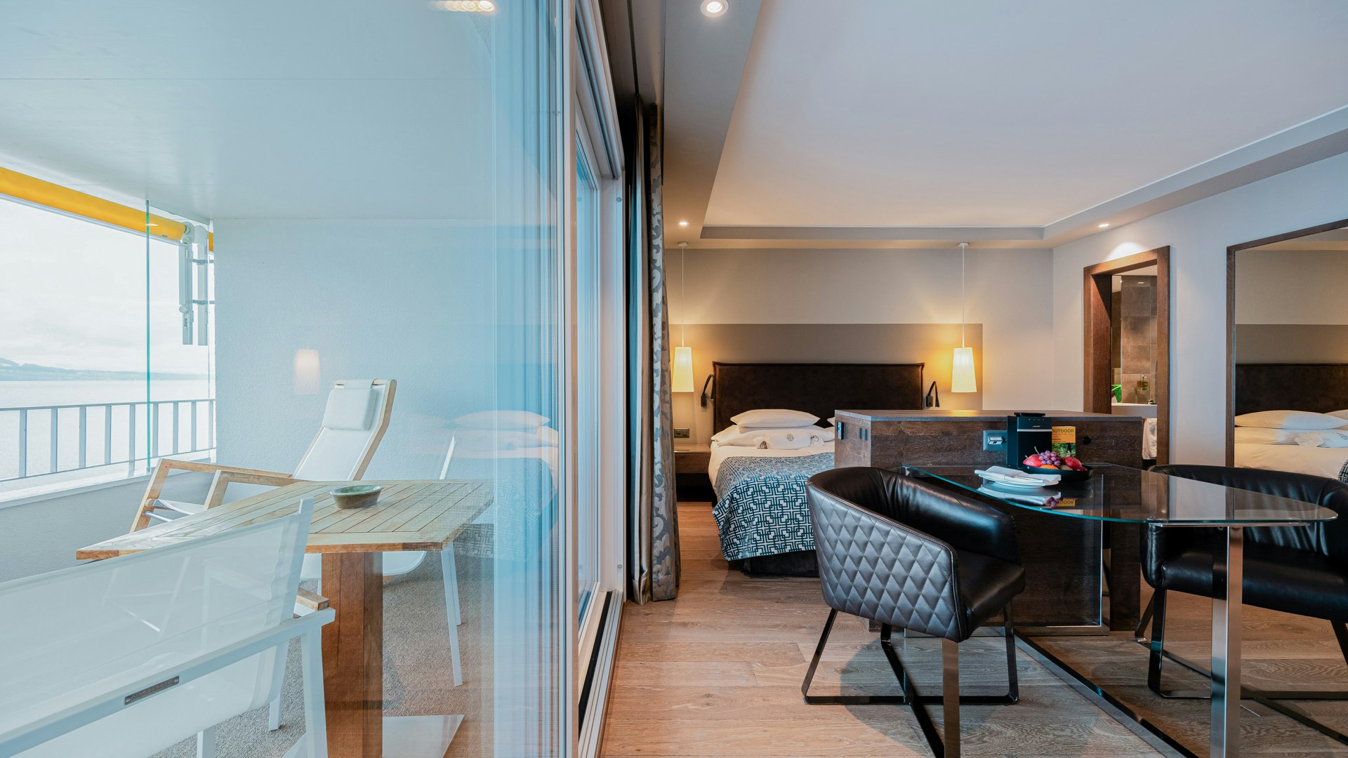 Eigerli Junior Suite in the Hotel BEATUS Left view through a window onto a balcony with lounger, right part of a room with double bed, armchair and glass table