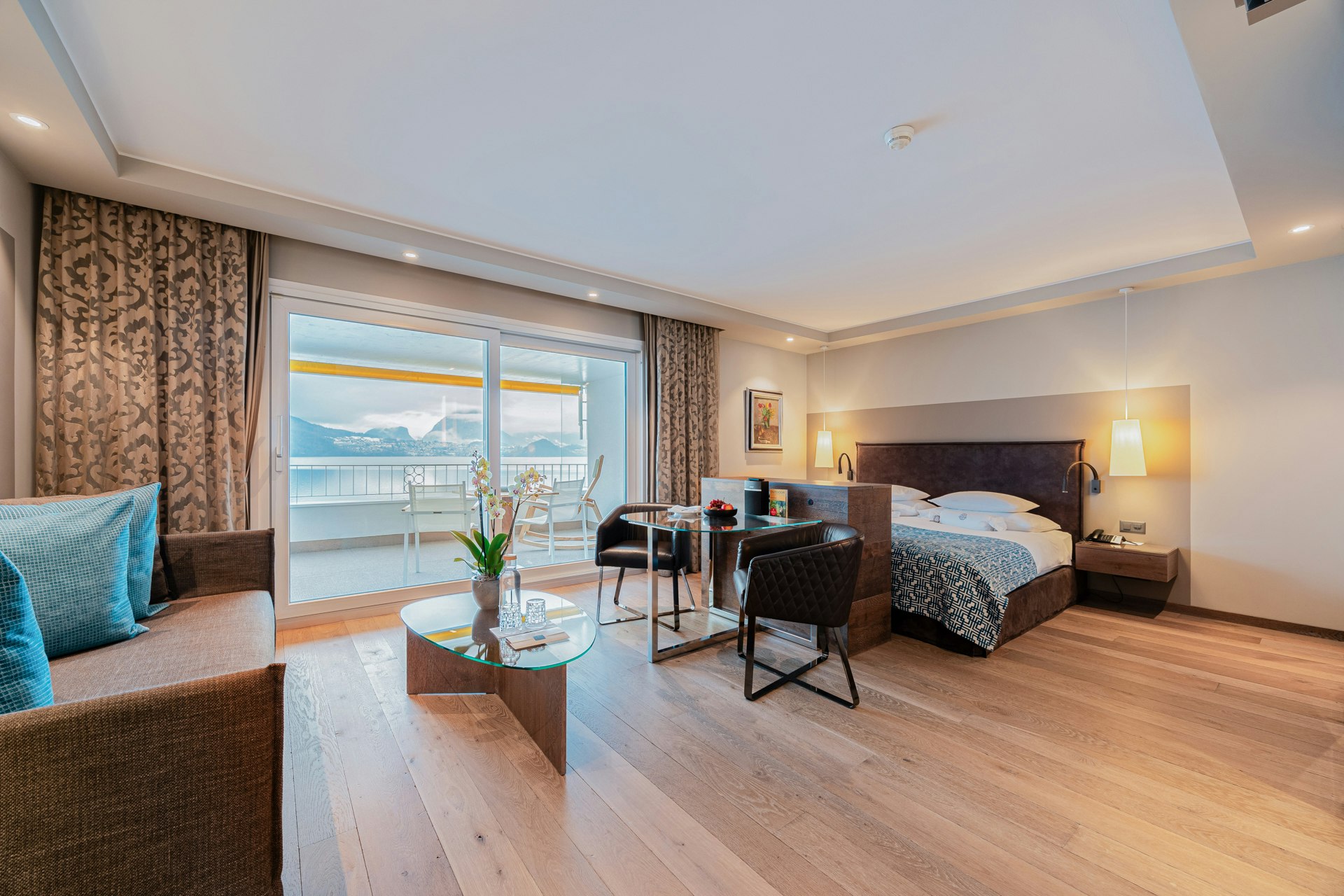 Wide-angle shot of a room with parquet flooring, sofa on the left, double bed on the right, glass table and armchair in the centre, large window and curtains