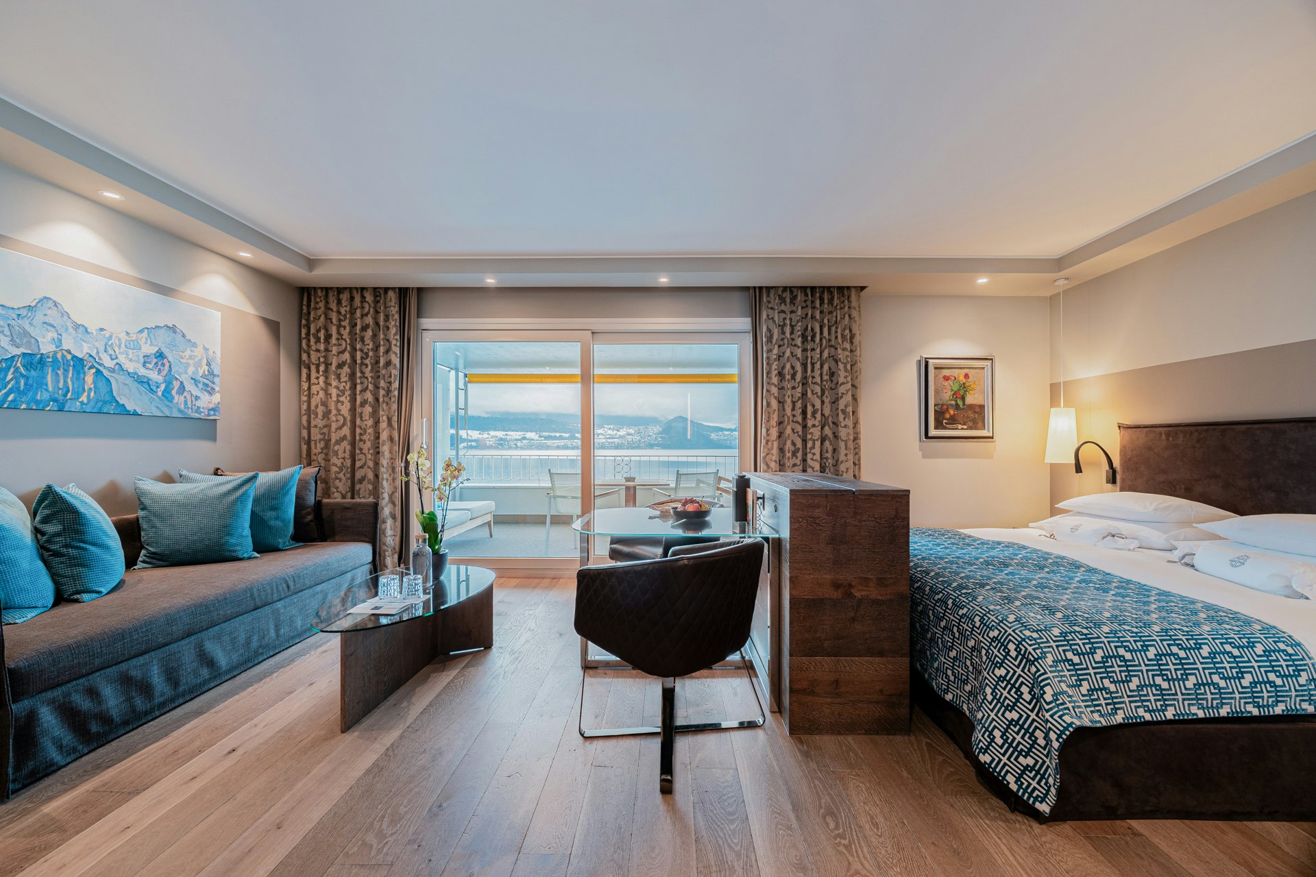 Room with parquet flooring, a sofa with cushions on the left, a picture of mountains above it, a glass table, an armchair and a half-height wardrobe in the centre, a double bed on the right, a large window at the very back