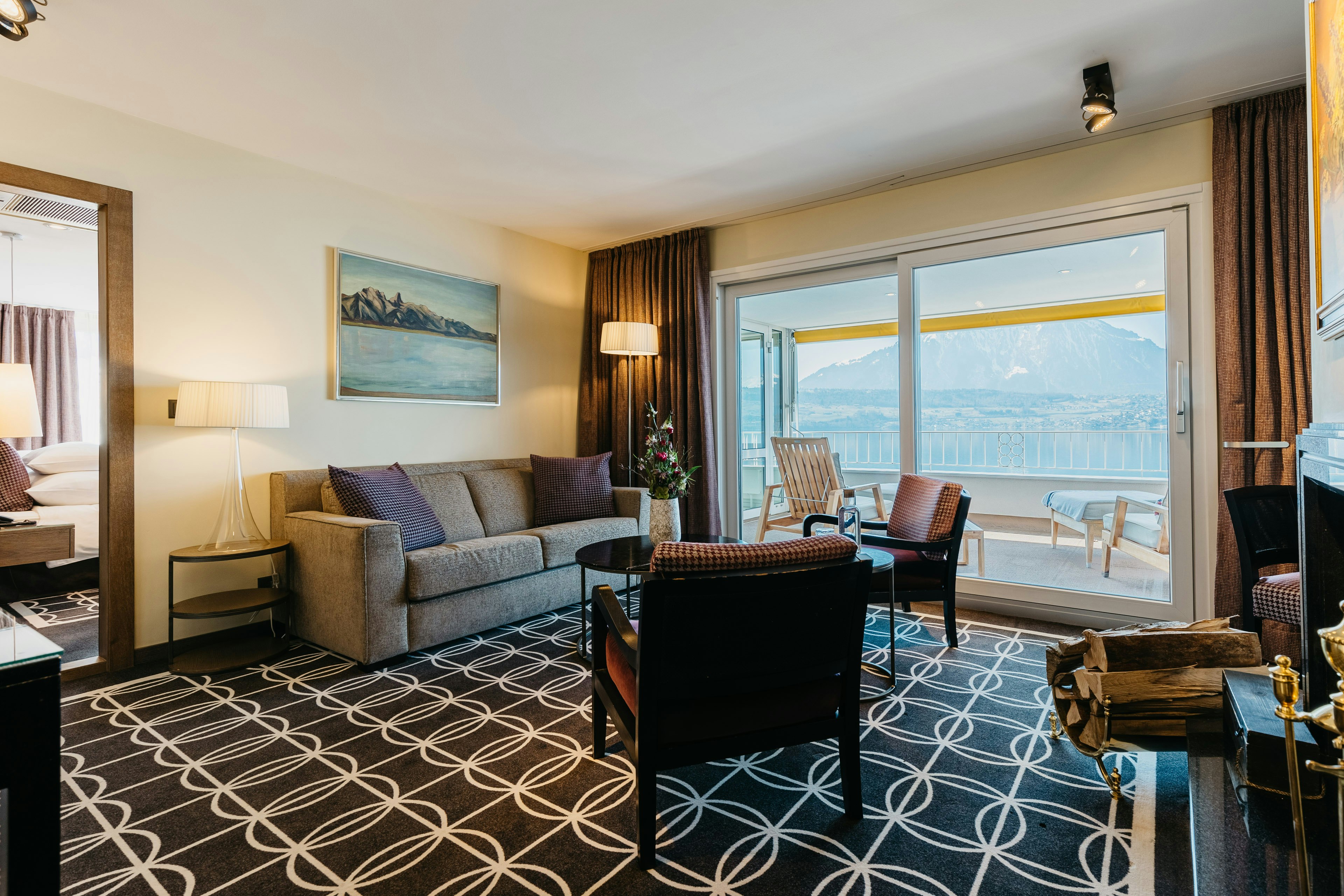 Zimmer mit Teppich, Sofa, Sesseln, kleinem Tisch und einem Panoramafenster mit Blick auf Balkon und Berg