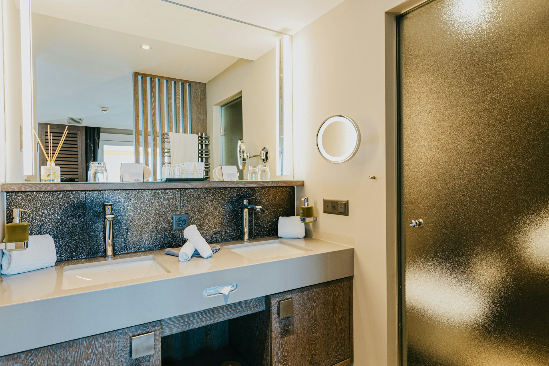 Badezimmer mit zwei Waschbecken, Spiegel, rechts eine halbtransparente Glastür, dahinter ein WC