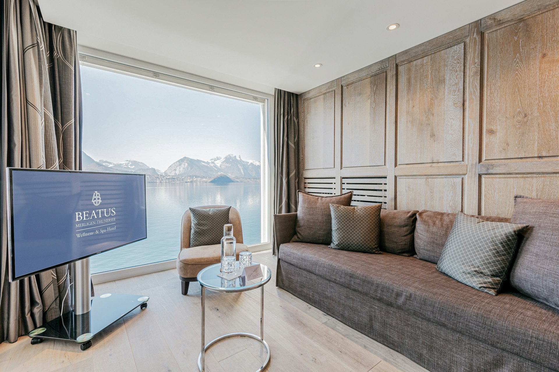 Blick auf ein Sofa mit Kissen, Sessel, Glastisch, Flachbildschirm, ein grosses Panoramafenster mit Blick auf See und Berge