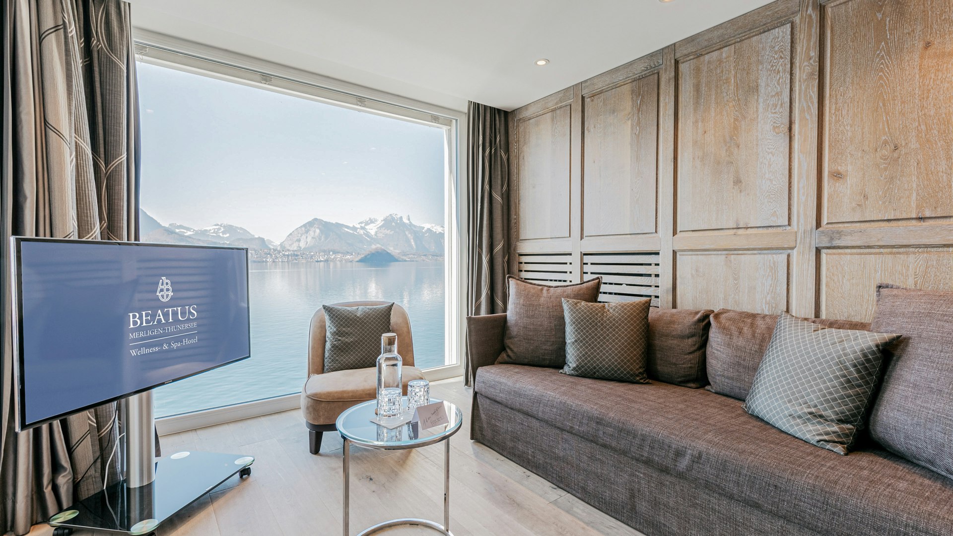 Senior Tower Suite in the Hotel BEATUS View of a sofa with cushions, armchair, glass table, flat screen, a large panoramic window with a view of the lake and mountains