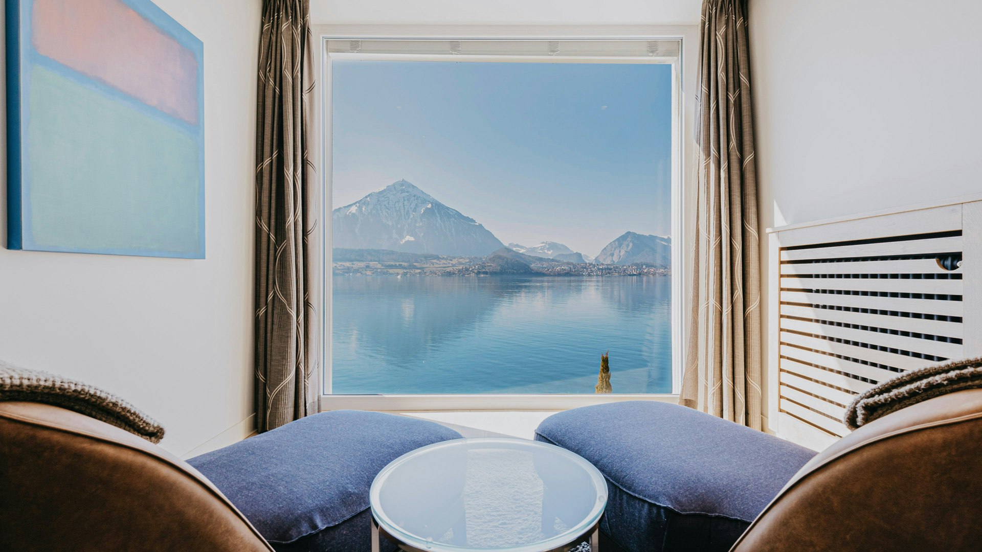 View from the tower suite in the Hotel BEATUS Erker with two pieces of upholstered furniture, a round glass table in between, an abstract painting on the left, a heater on the right, all in front of a panoramic window with a view of the lake and mountains