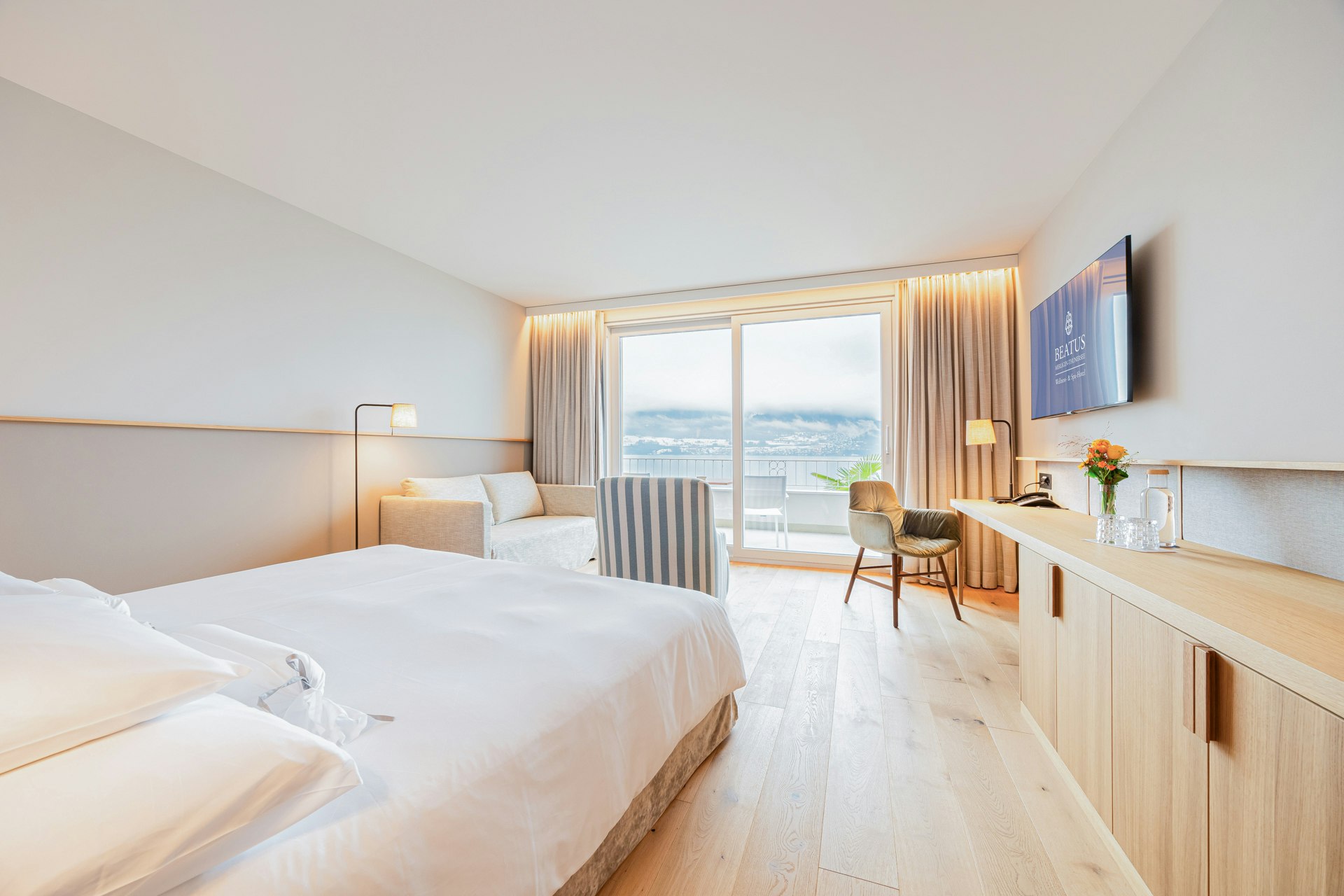 Links ein Doppelbett, rechts ein hellbrauner Holzkorpus, hinten ein Sofa, Polstersessel, ein Stuhl und an der Wand ein Flachbildschirm, ganz hinten ein Panoramafenster