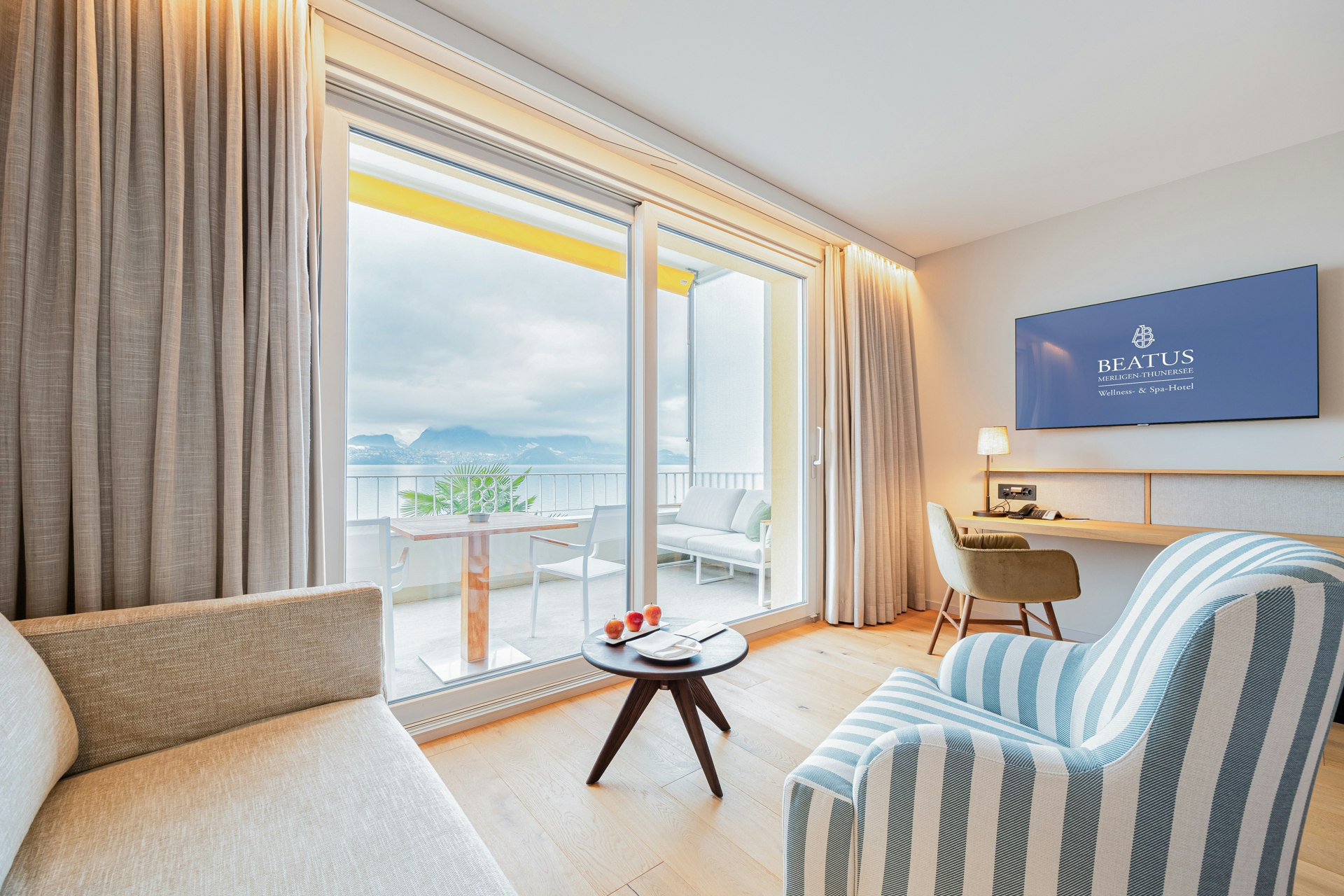 Zimmer mit Sofa, gestreiftem Polstersessel, rundem Holztisch, hinten ein Stuhl, Tisch und darüber ein Flachbildschirm, links ein Panoramafenster mit Balkon