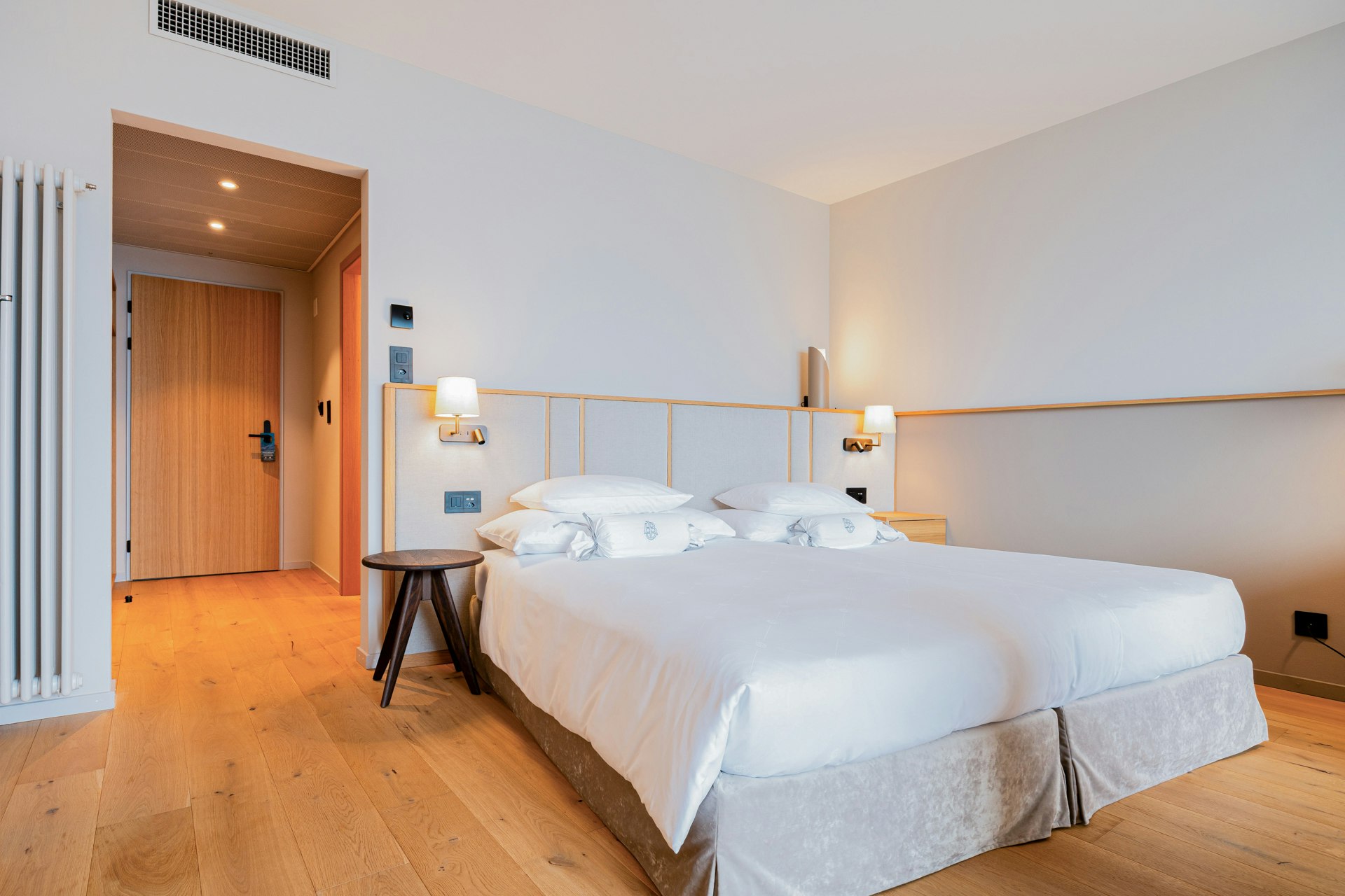 View of a double bed, a stool next to it, a corridor at the back left, parquet flooring