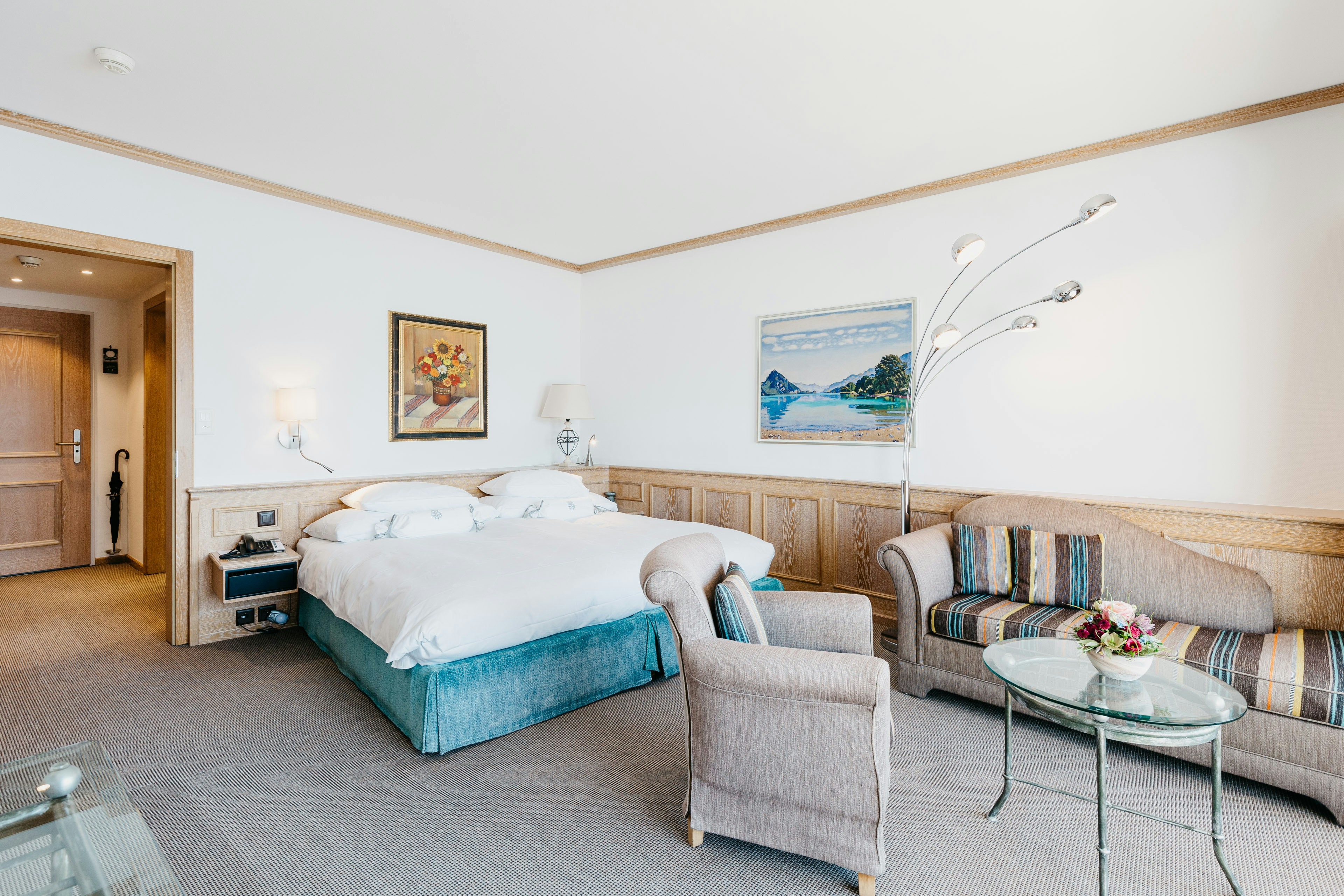 Zimmer mit Polstermöbel, dahinter ein Doppelbett, halbhohe Holzvertäfelung, Stillleben an den Wänden
