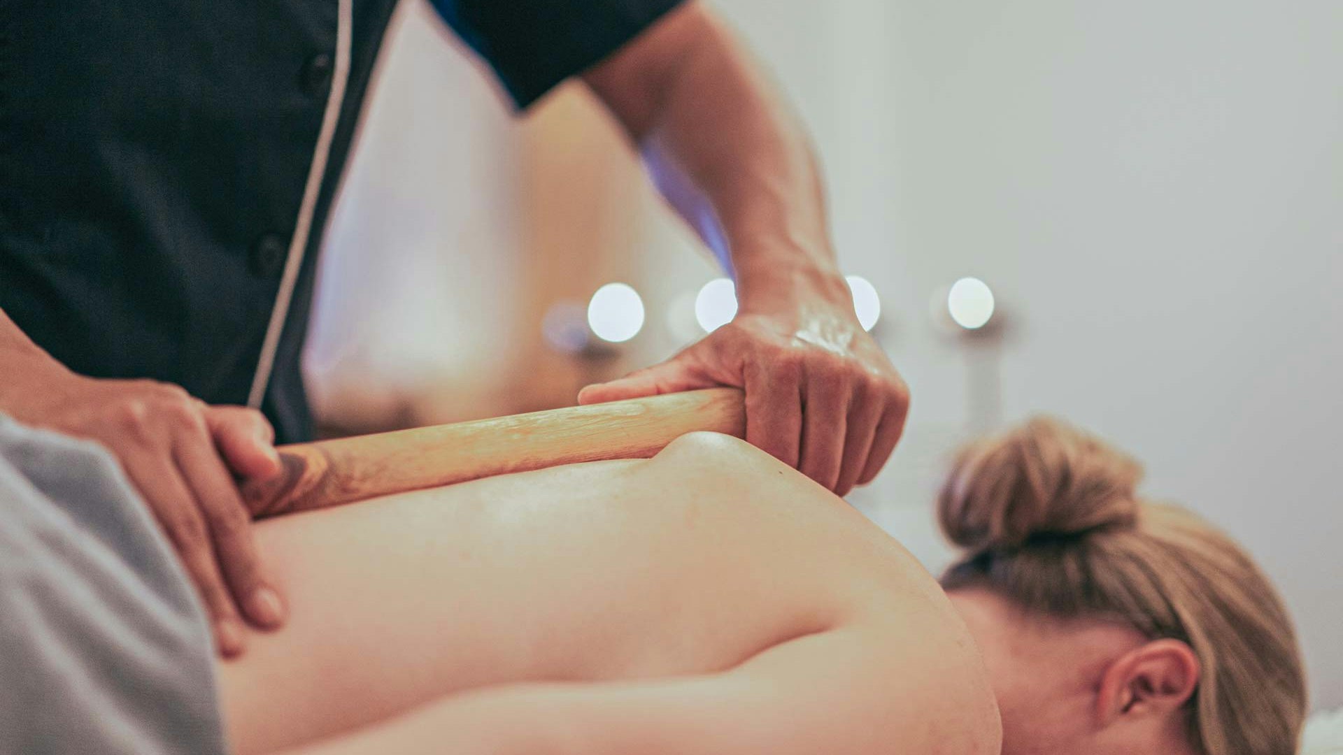 Zirben-Vital-Massage im Hotel BEATUS Eine Frau liegt auf dem Bauch auf einer Massageliege und wird mit einem Zirbenstock massiert