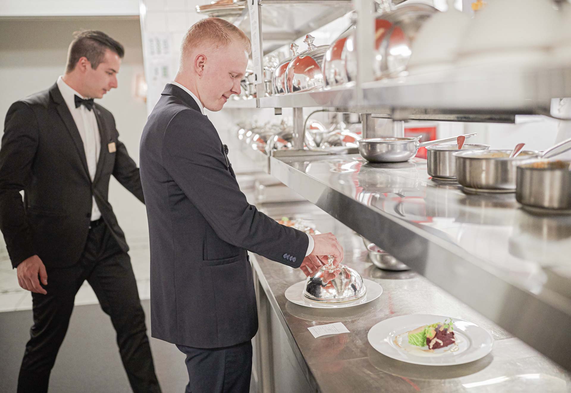 Two men in dark suits, white shirts and with bow ties around their necks take plates of food from a canteen kitchen