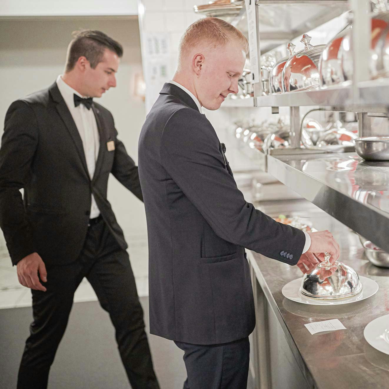Personnel de cuisine à l'hôtel BEATUS Deux hommes en costume sombre, chemise blanche et nœud papillon autour du cou prennent des assiettes de nourriture dans une grande cuisine.