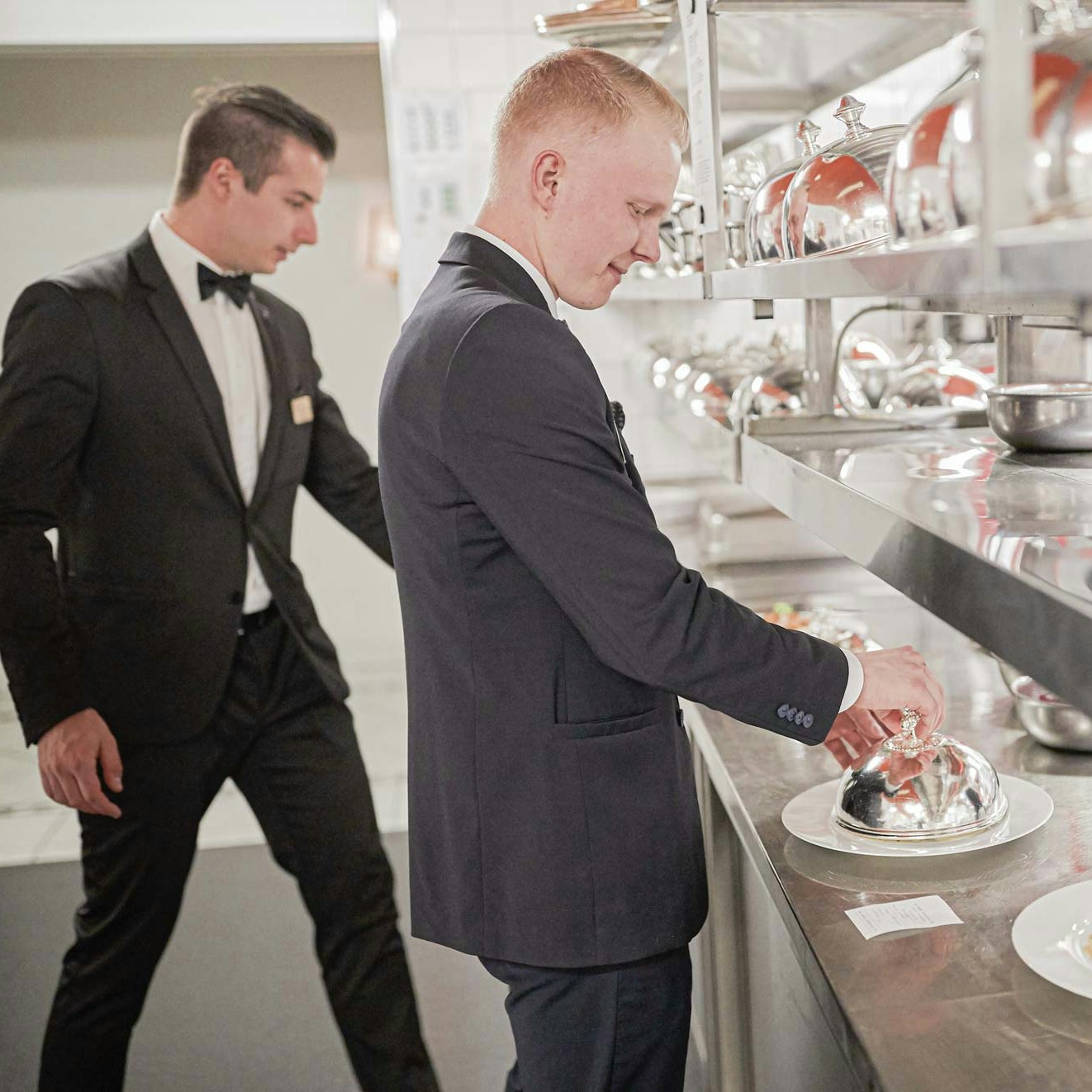 Küchenpersonal im Hotel BEATUS Zwei Männer im dunklen Anzug, weissen Hemd und mit Fliege um den Hals nehmen Teller mit Speisen aus einer Grossküche