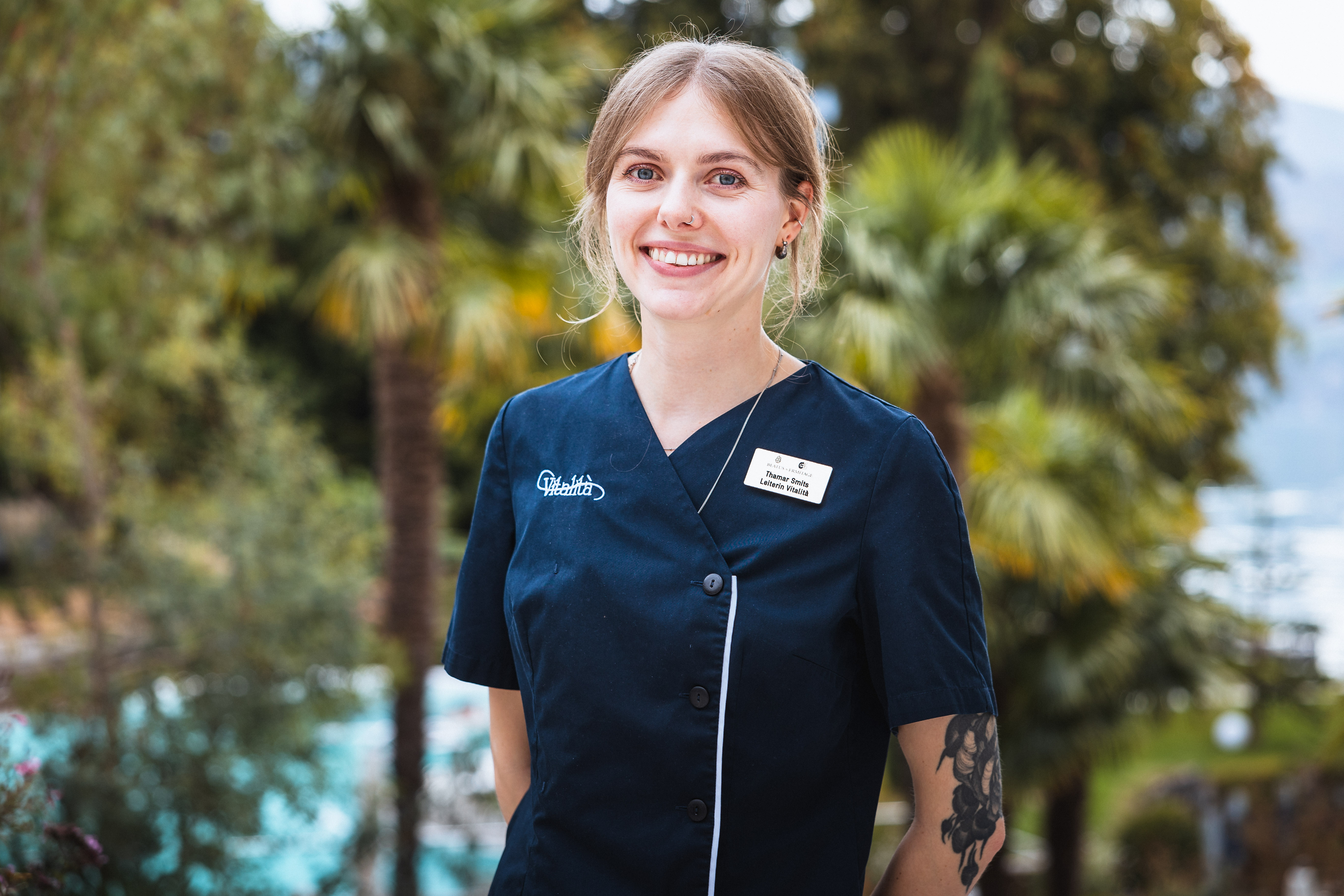 Lächelnde junge Frau in dunkler Uniform mit Tattoos am Arm, im Hintergrund unscharf Palmen