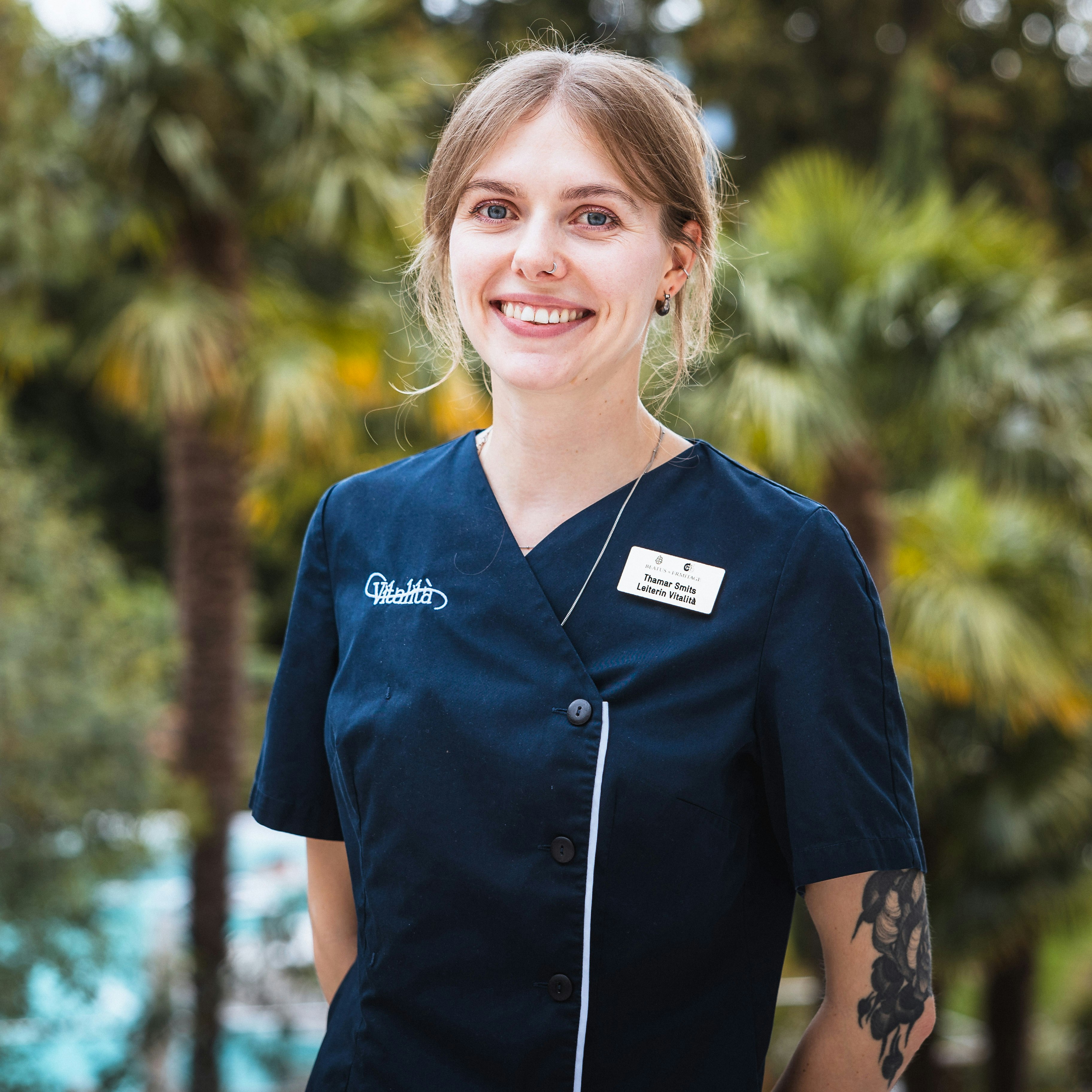 Thamar Smits, directrice de Vitalità Jeune femme souriante en uniforme sombre avec des tatouages sur le bras, palmiers flous en arrière-plan
