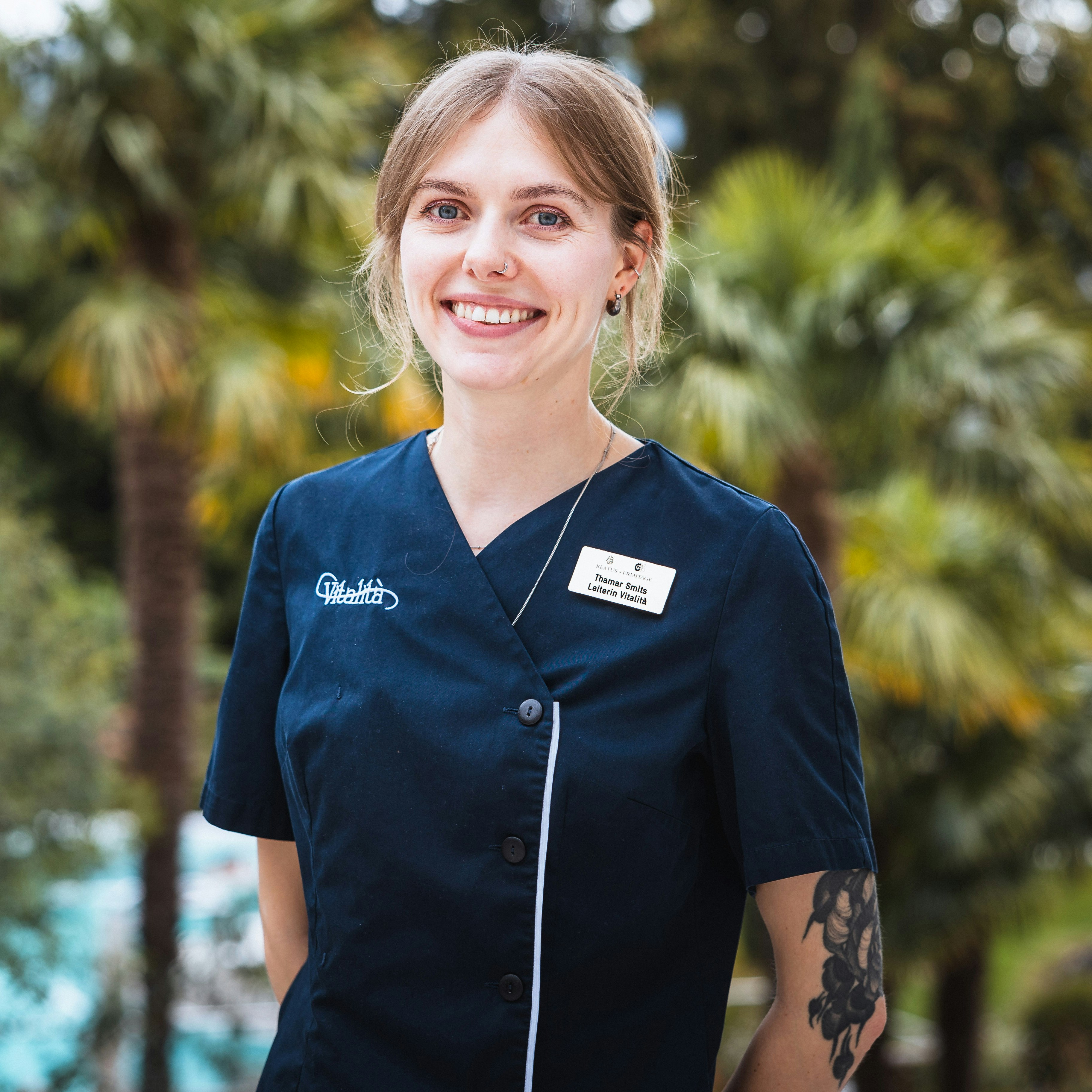 Thamar Smits, Leiterin Vitalità Lächelnde junge Frau in dunkler Uniform mit Tattoos am Arm, im Hintergrund unscharf Palmen