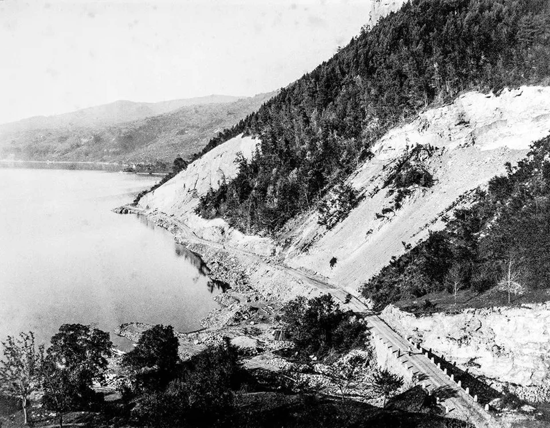 Road construction between Gunten, Merligen and Beatenbucht, 1837 A black and white photo with a lake on the left and a steep mountain slope on the right from which a road is being built