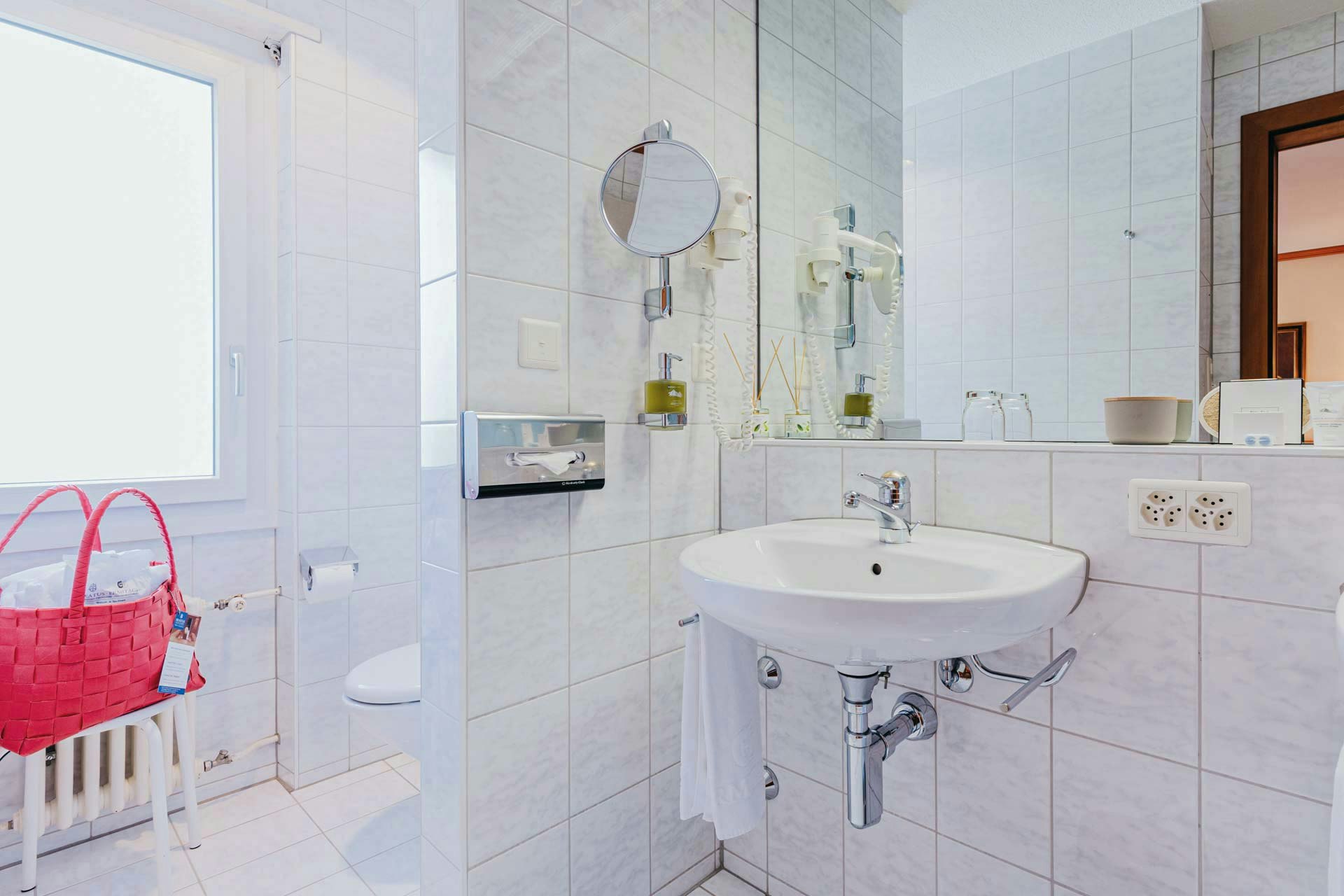 On the left a niche with WC, in front of it a red bathing bag on a stool, on the right a washbasin with a large mirror above it