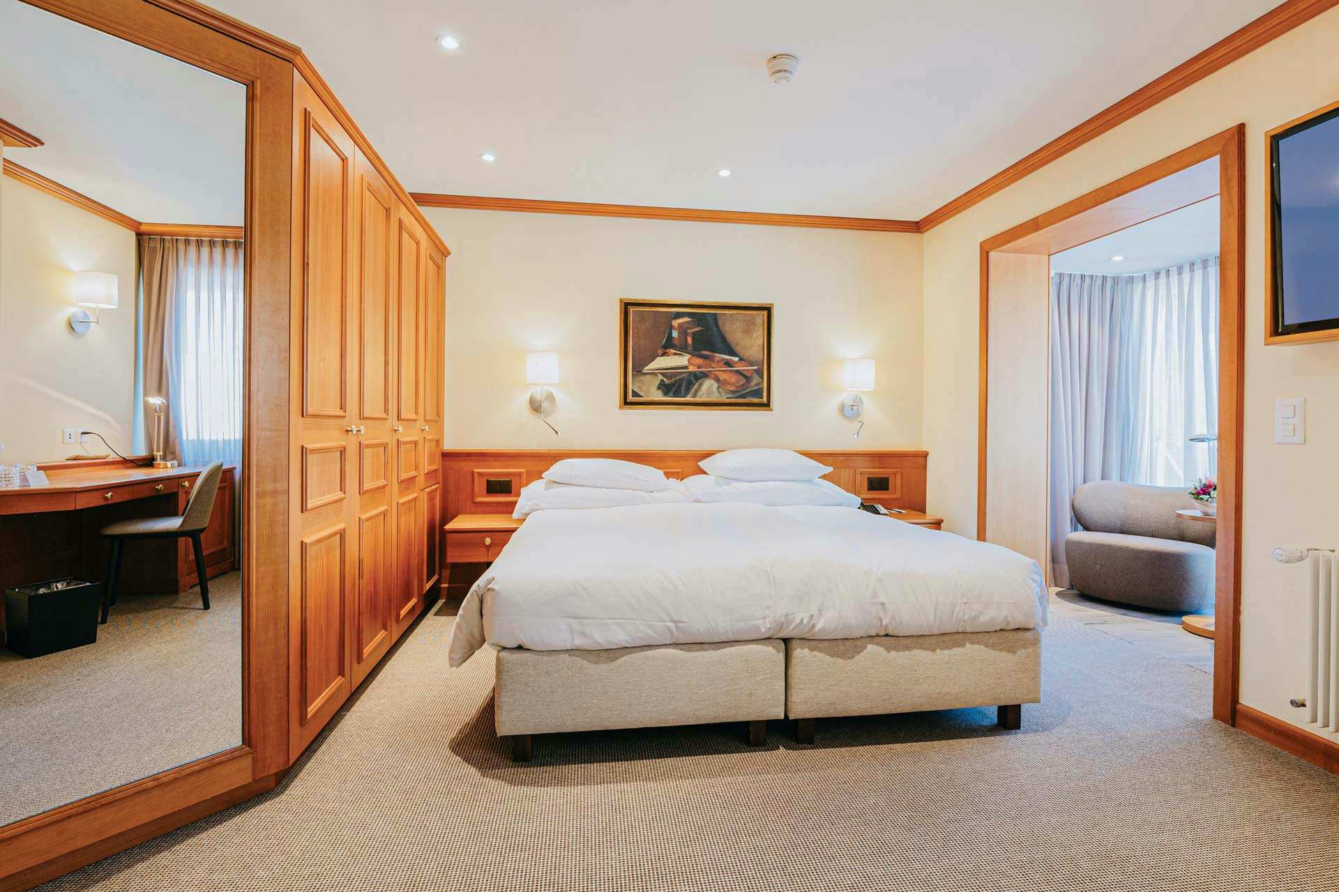 Room with carpeted floor and wall unit, in the centre a double bed, on the right you look into a room with an upholstered armchair and a large window