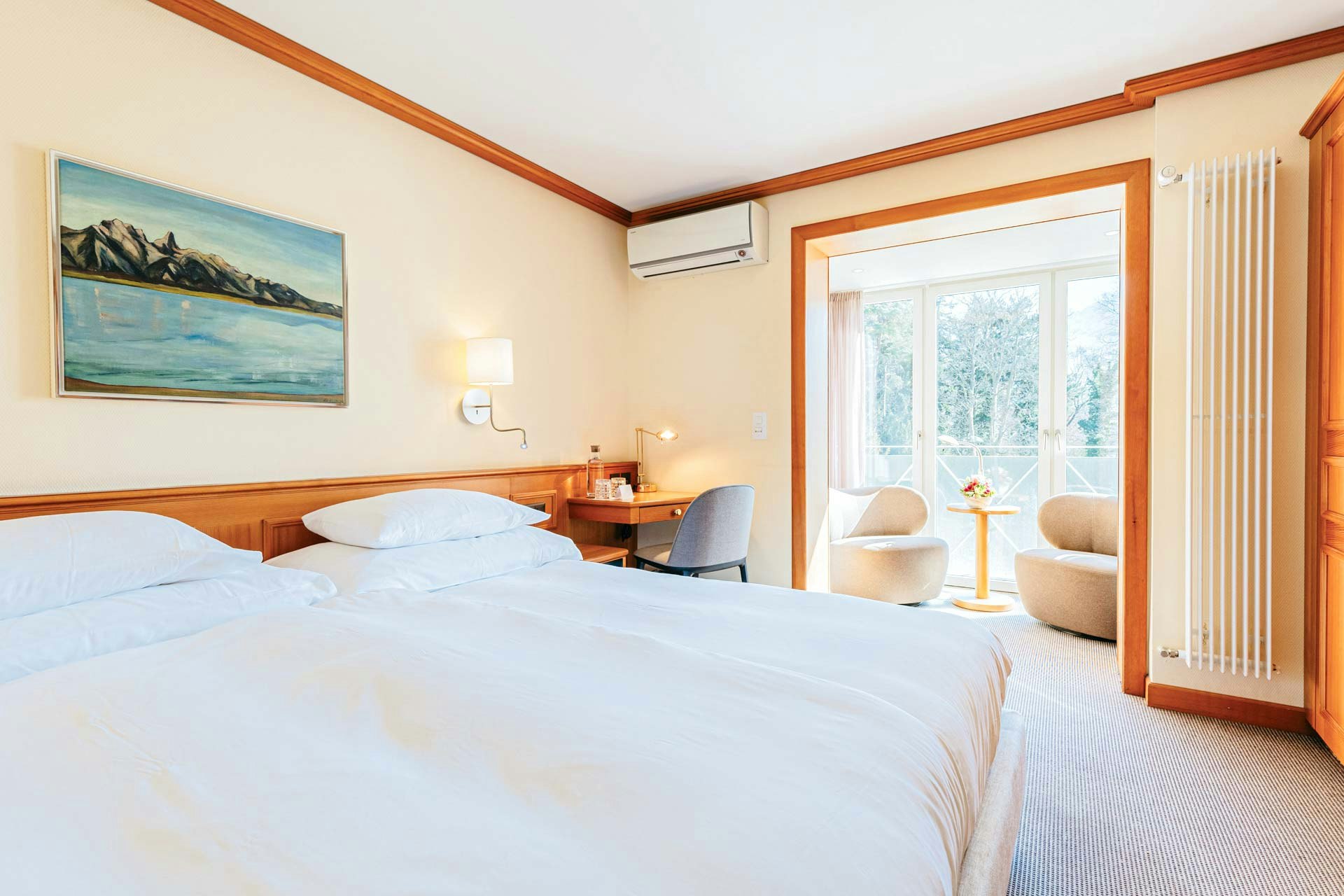 Zimmer mit Teppichboden, links ein Doppelbett mit einem Landschaftsbild, hinten blickt man in einen Raum mit Polstersesseln und einem grossen Fenster