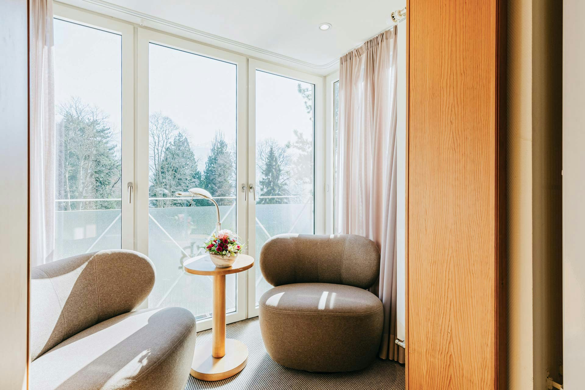 View into a sunny little room with floor-to-ceiling windows, a sofa in front, a round table with flowers and an upholstered armchair