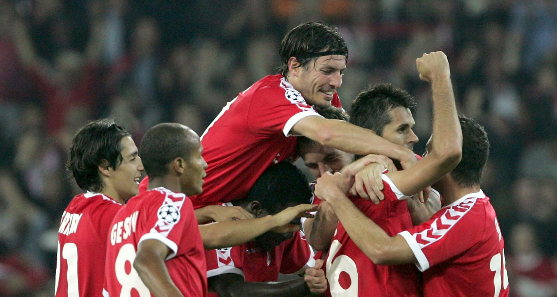 Cinq joueurs de football en tenue rouge, l'un sautant sur l'autre qui lève le bras