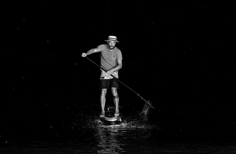 Schwarz-weiss Foto eines Mannes auf einem SUP bei Nacht, mit einem flachen Hut und einem gestreiften T-Shirt bekleidet