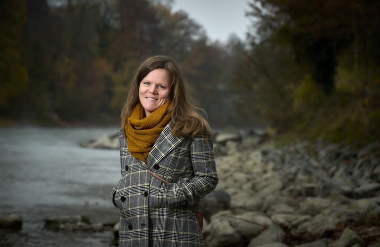 Erika Kneubühl, Course Leader Retreats Woman in a chequered coat and scarf stands in front of a dark river landscape