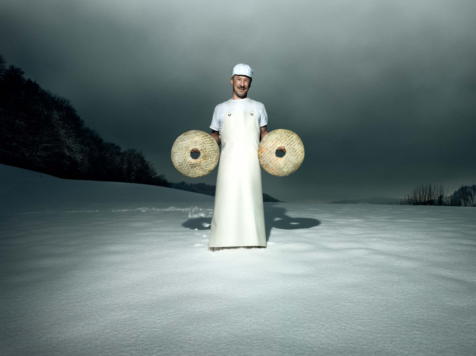 A man stands in the snow wearing a long white apron, holding a wheel of