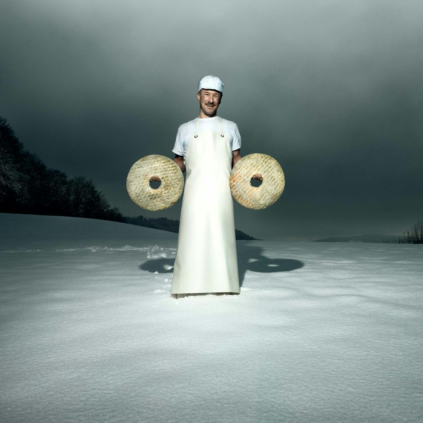 Willi Schmid, Käser Ein Mann steht in langer weisser Schürze im Schnee, links und rechts einen Laib Käse in der Hand