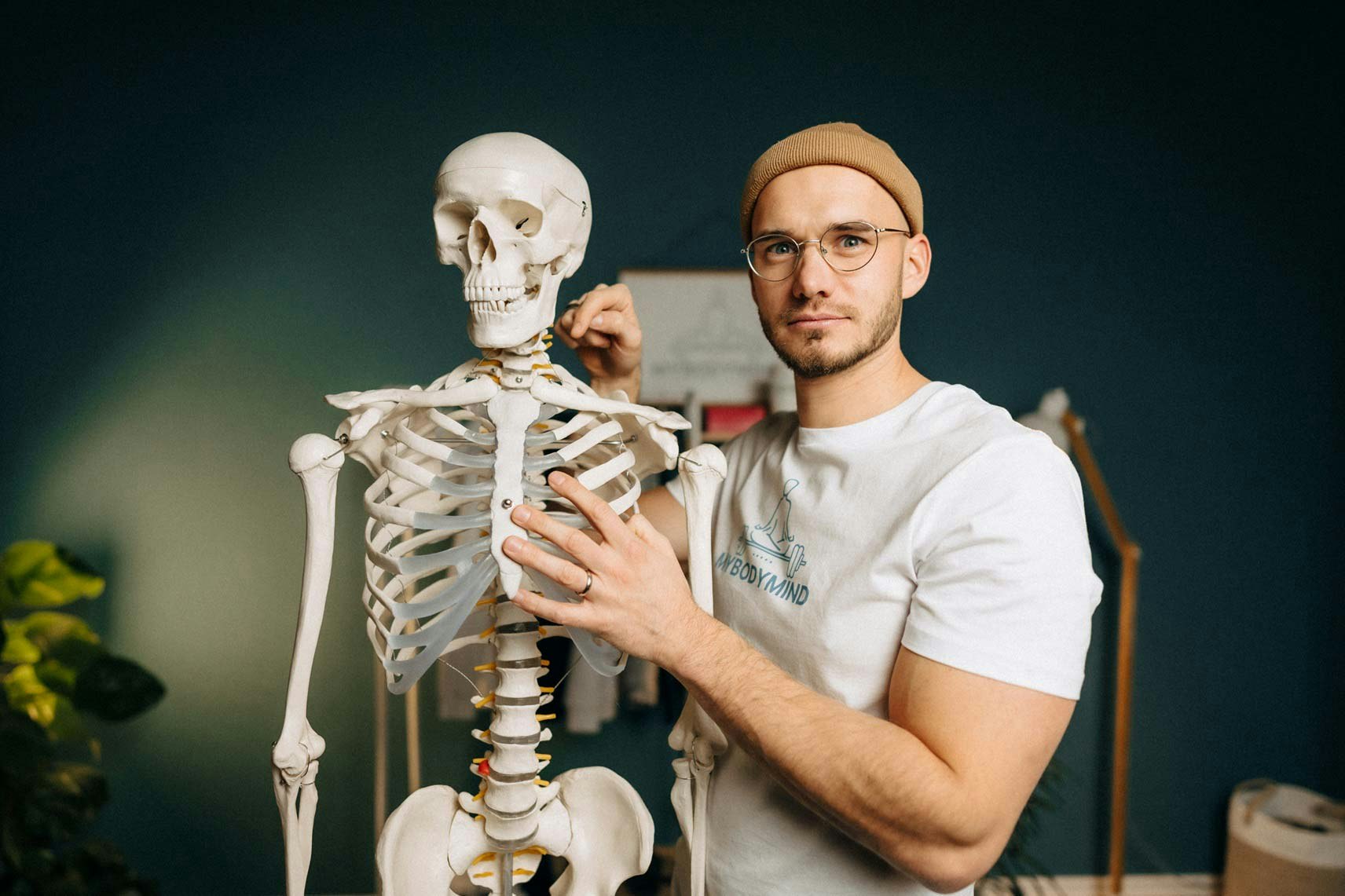 Junger Mann mit Brille und Bart, eine Mütze am Kopf, steht neben einem Modellskelett