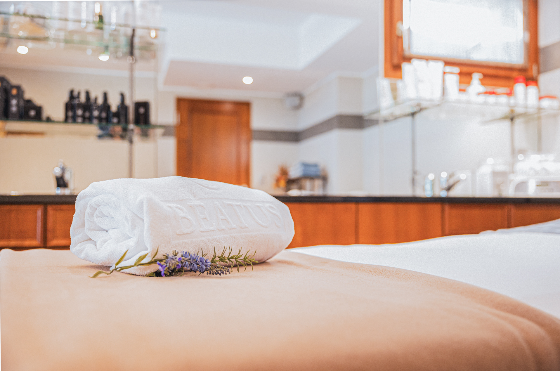 Une serviette enroulée est posée sur une table de massage, une branche de lavande est placée devant elle, et différents produits de soins sont posés sur une armoire en arrière-plan.