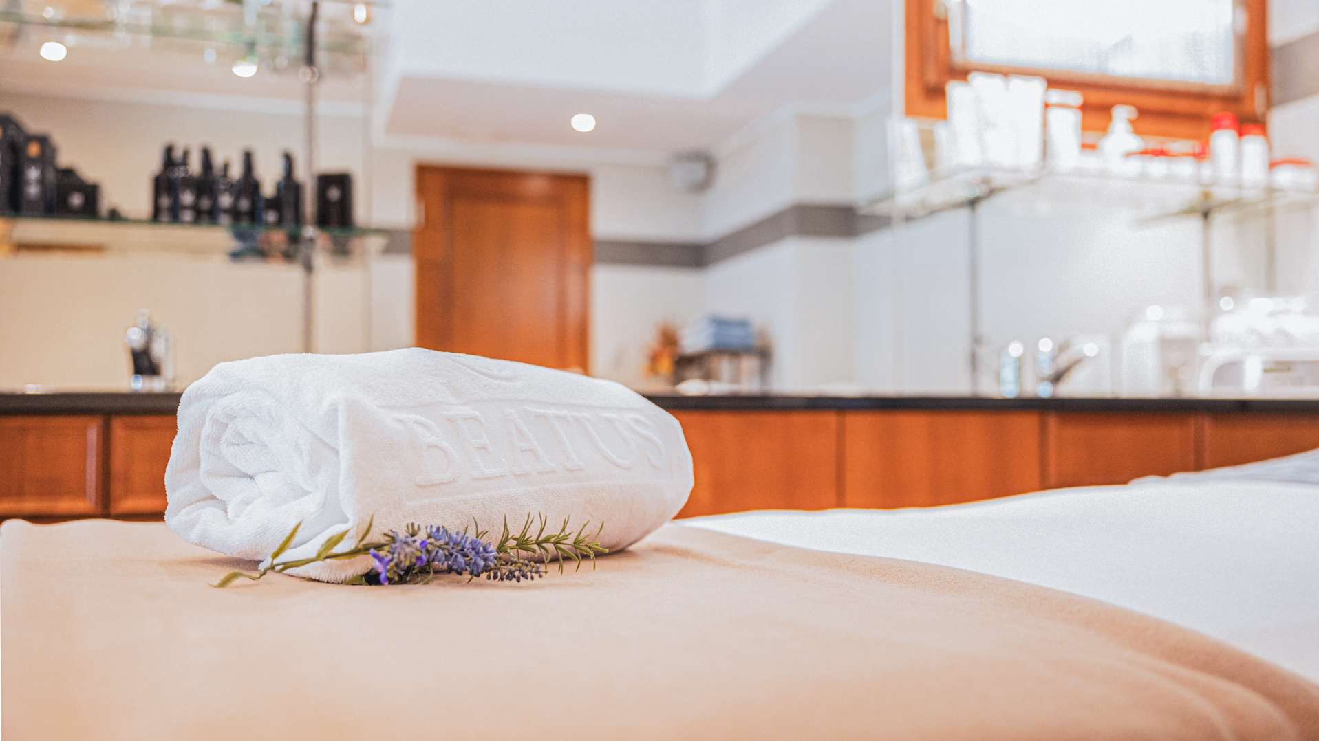 Soins de beauté à l'hôtel BEATUS Une serviette enroulée est posée sur une table de massage, une branche de lavande est placée devant elle, et différents produits de soins sont posés sur une armoire en arrière-plan.