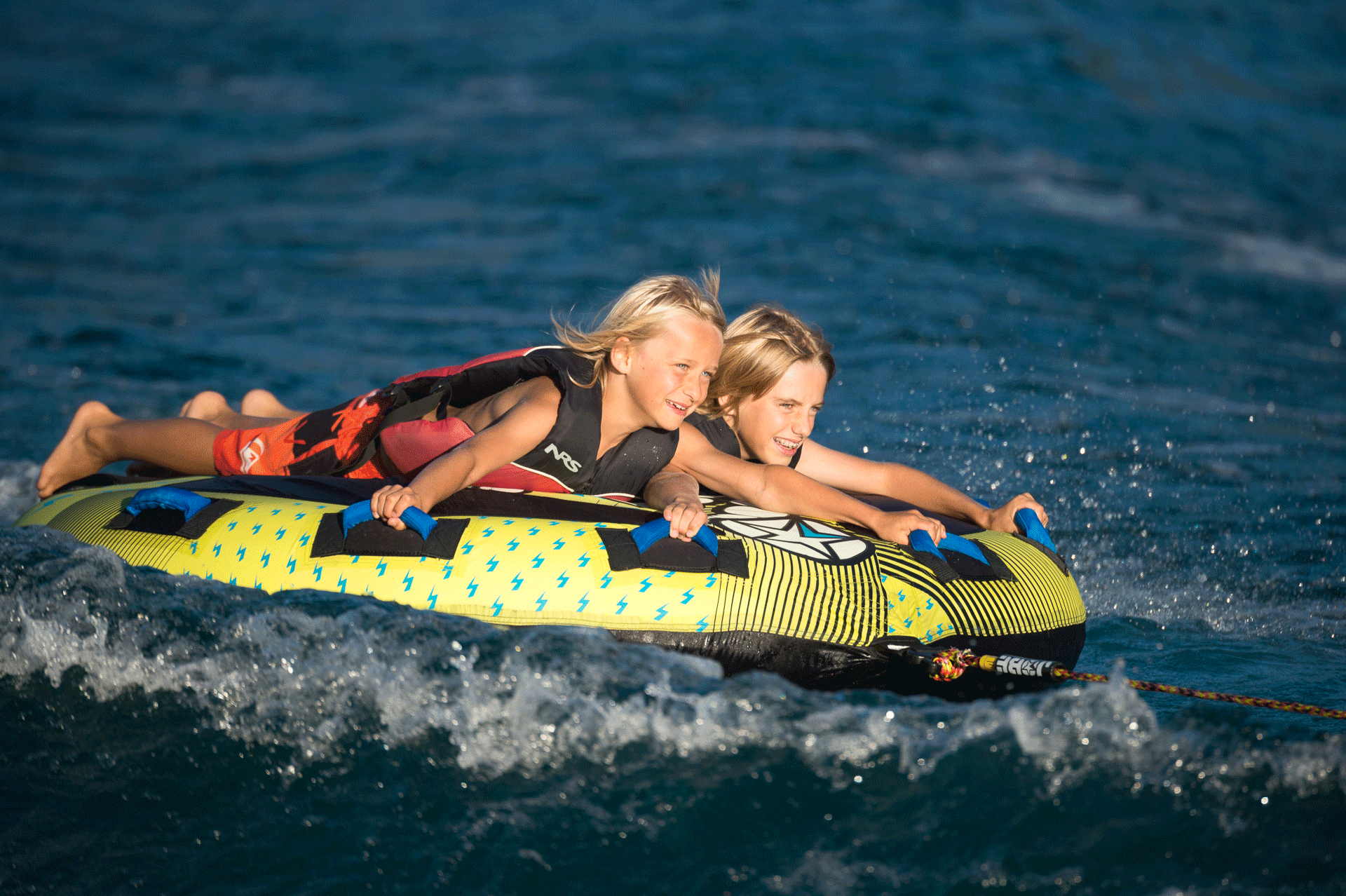 Zwei lachende Kinder liegen auf einem grossen Gummireifen im Wasser und lassen sich von einem Boot ziehen