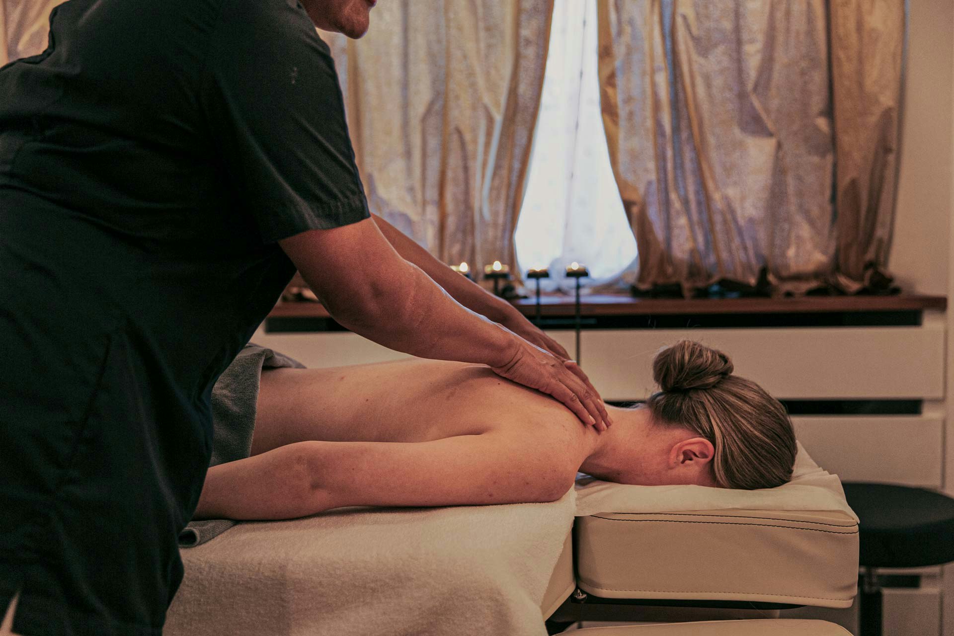 A woman lies on her stomach on a massage table and is massaged on the shoulders by another woman
