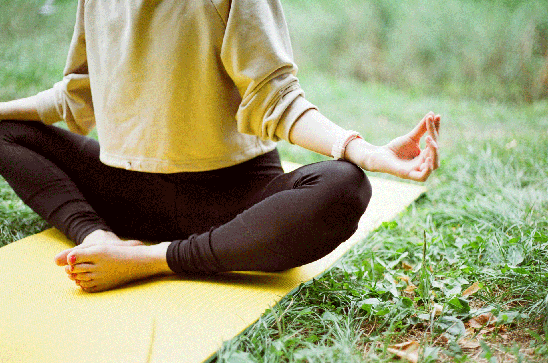 Woman in yellow jumper sitting on yellow yoga mat in greenery, fingers in mudra position
