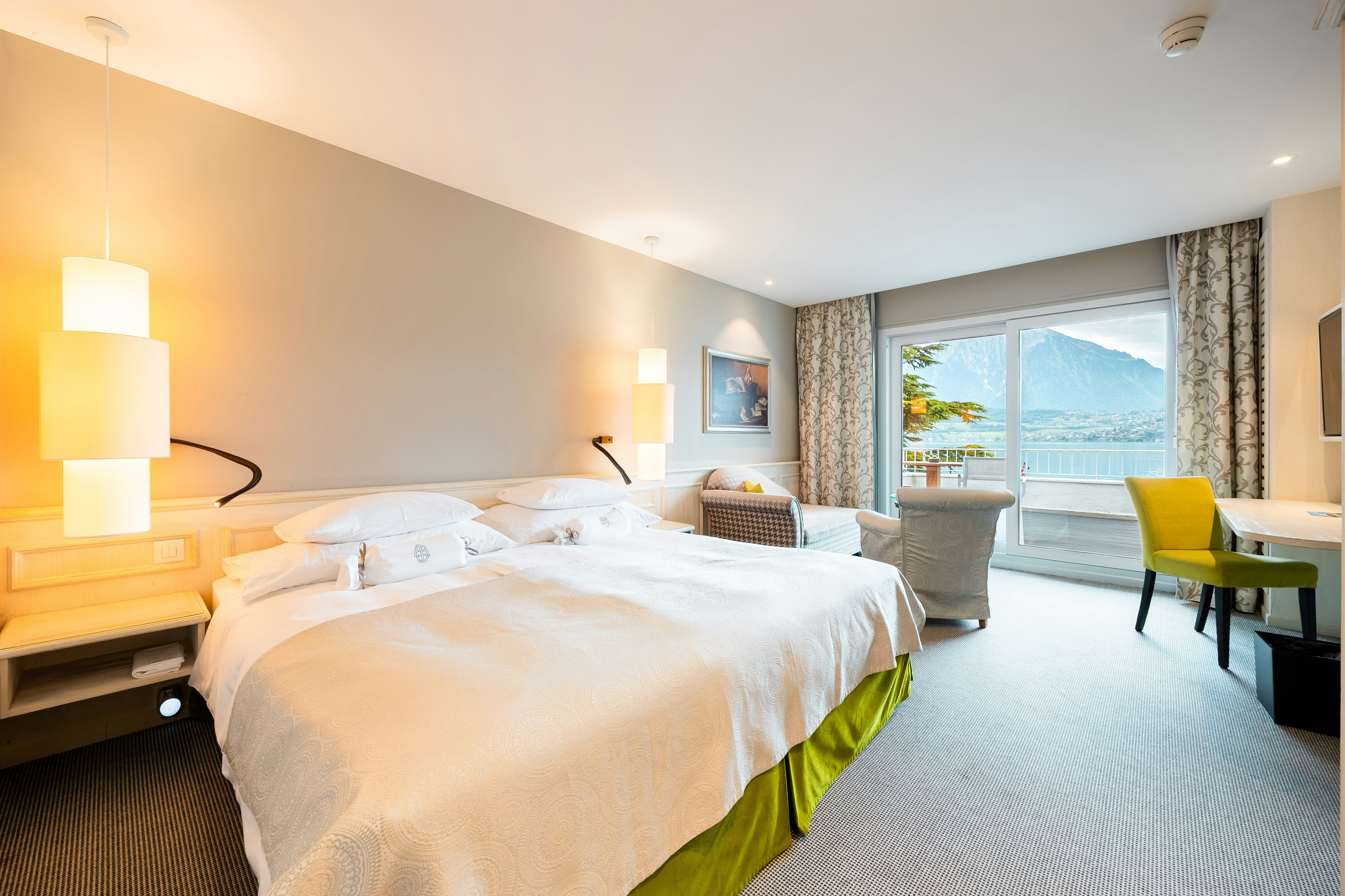 Zimmer mit Doppelbett, Hängeleuchte, dahinter Sofa, Polstersessel, Glastisch, rechts an der Wand ein Flachbildschirm, darunter ein Stuhl vor einem Klapptisch, hinten Panoramafenster mit Seeblick,