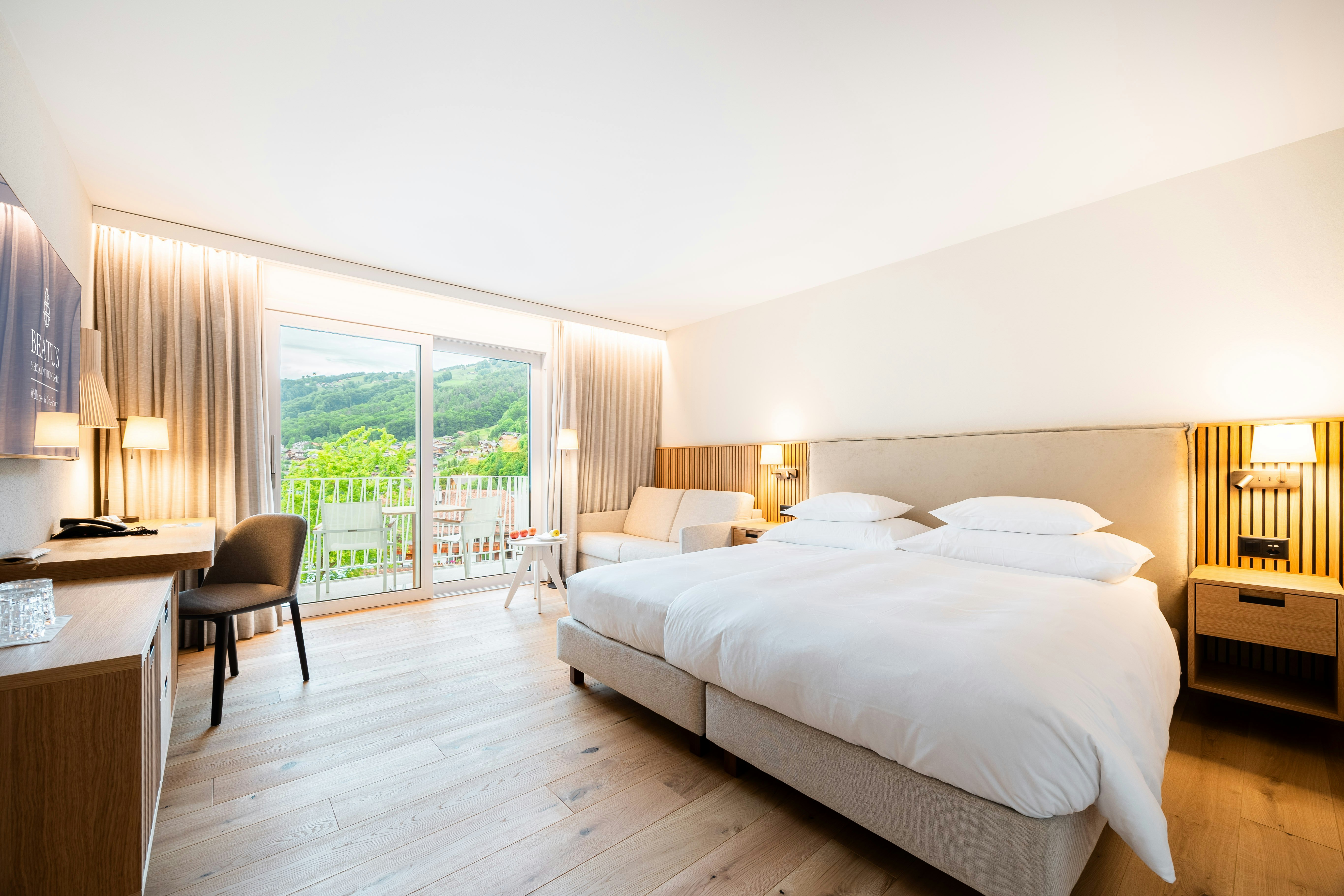Doppelbett in der Mitte des Bildes, Parkettboden, links Ablagefläche, Pult, Fernseher und Stuhl, und Balkon mit zwei Stühlen und kleinem Tisch