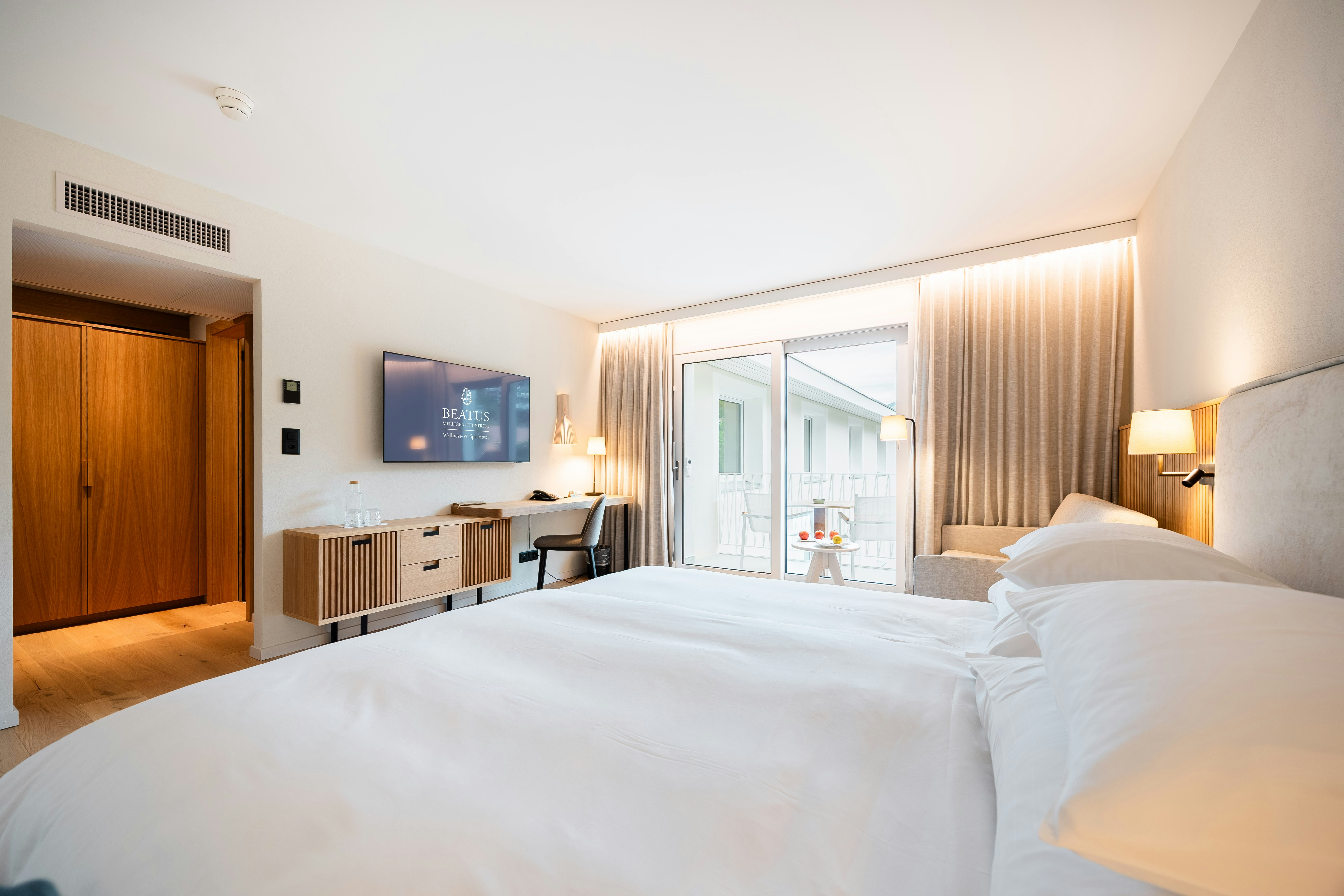 In the foreground, a double bed; in the centre of the image, a television, a storage surface and a desk; on the right, a balcony with a view of Sigriswil, the road, and a limited lake view.