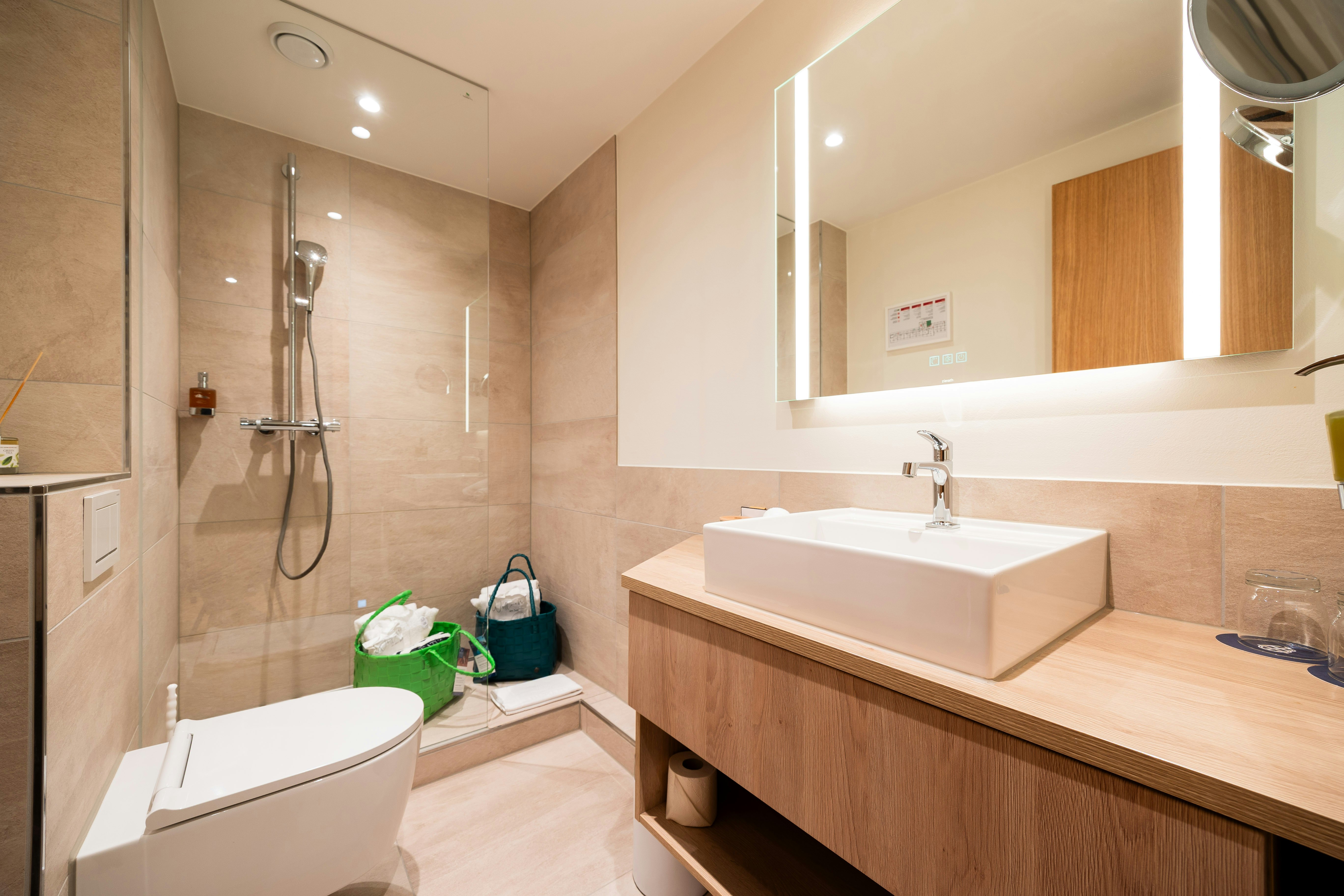 Badezimmer in Sandfarben im Vordergrund links WC, hinten eine Dusche mit kleinem Absatz und rechts ein Lavabo mit Spiegel