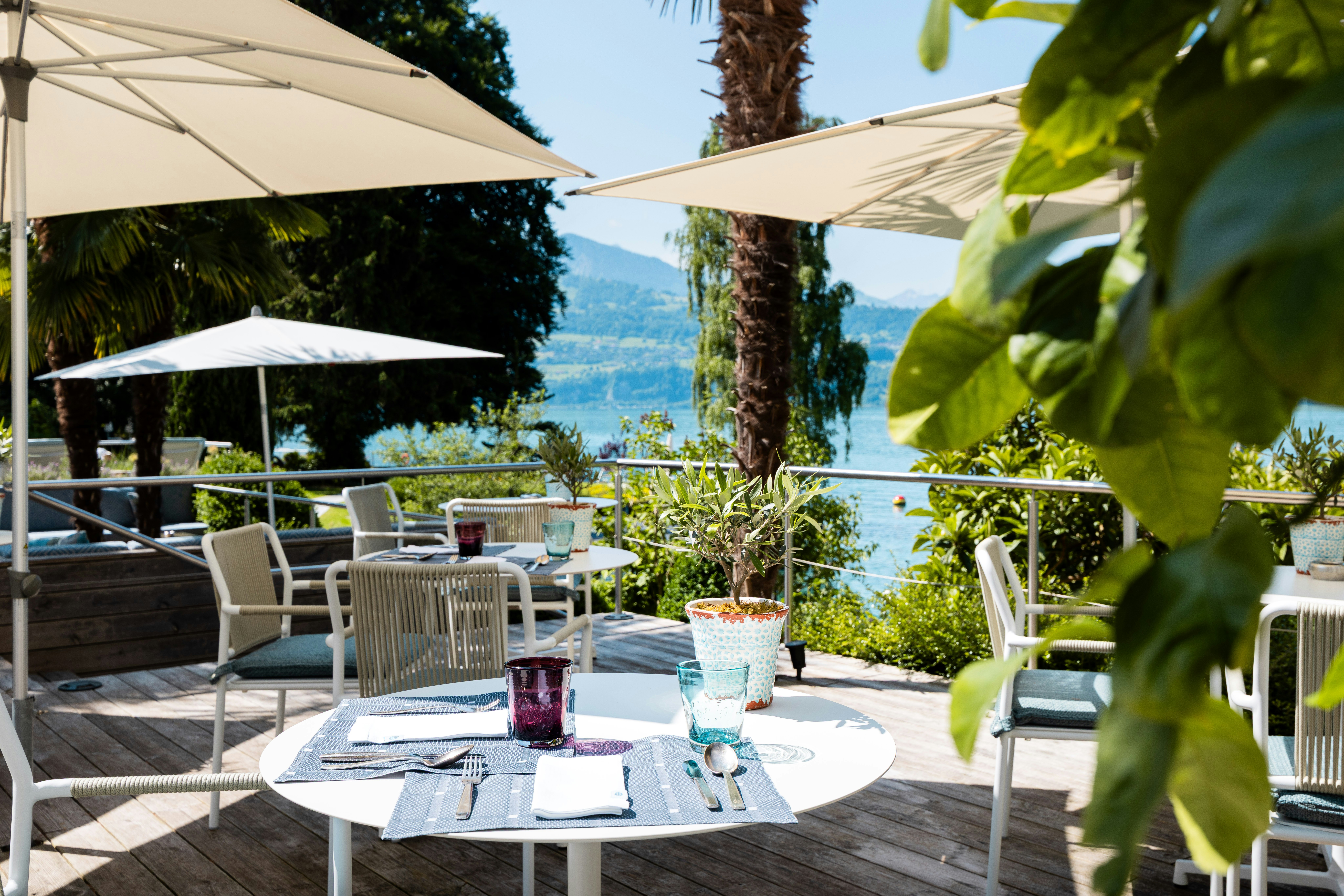 A set table in the foreground with placemats, glasses, cutlery, and floral decoration. In the distance, Lake Thun shimmers in the background
