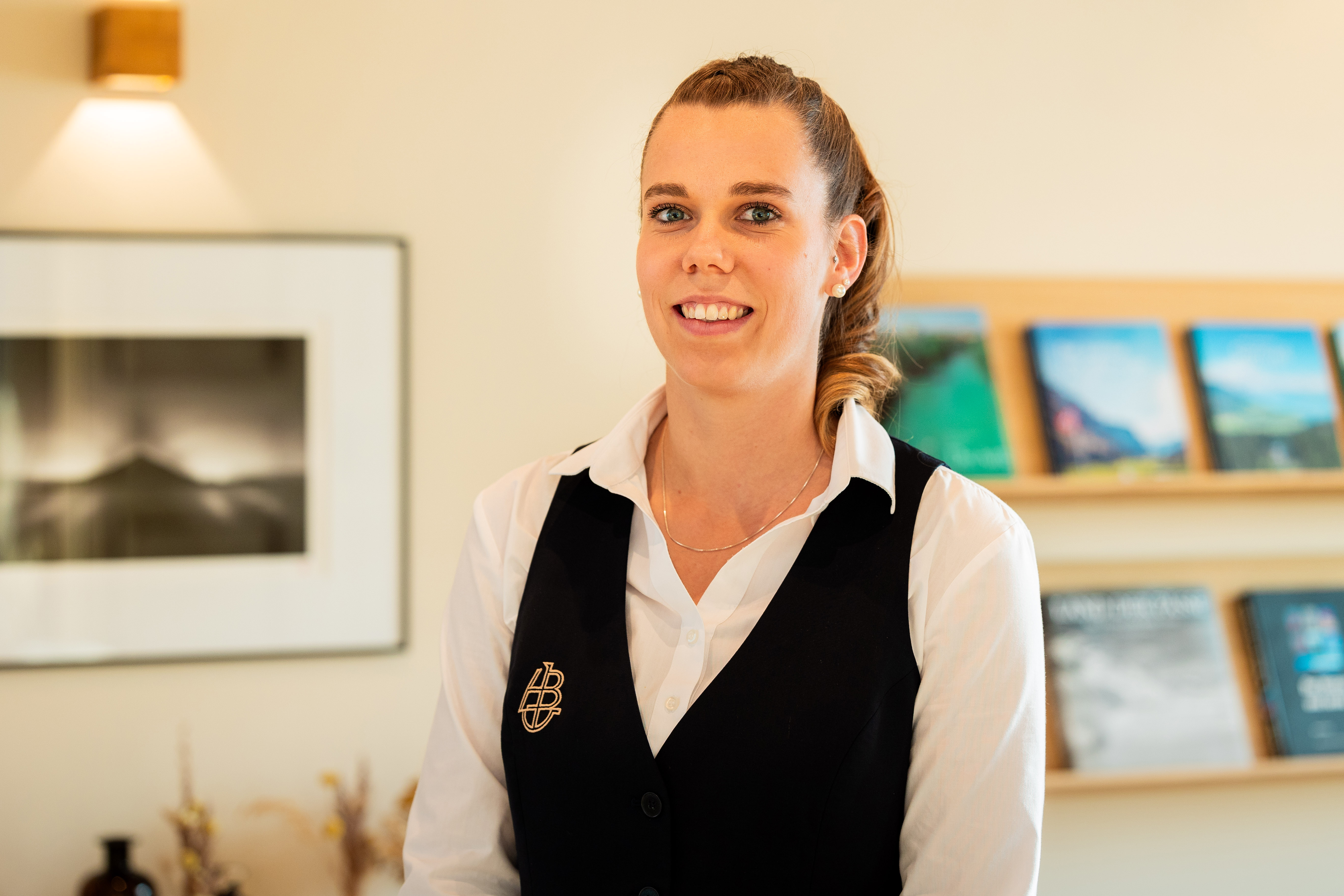 Lächelnde Frau mit weisser Bluse und schwarzem Gilet vor einem Regal mit Magazinen und einem gerahmten Foto