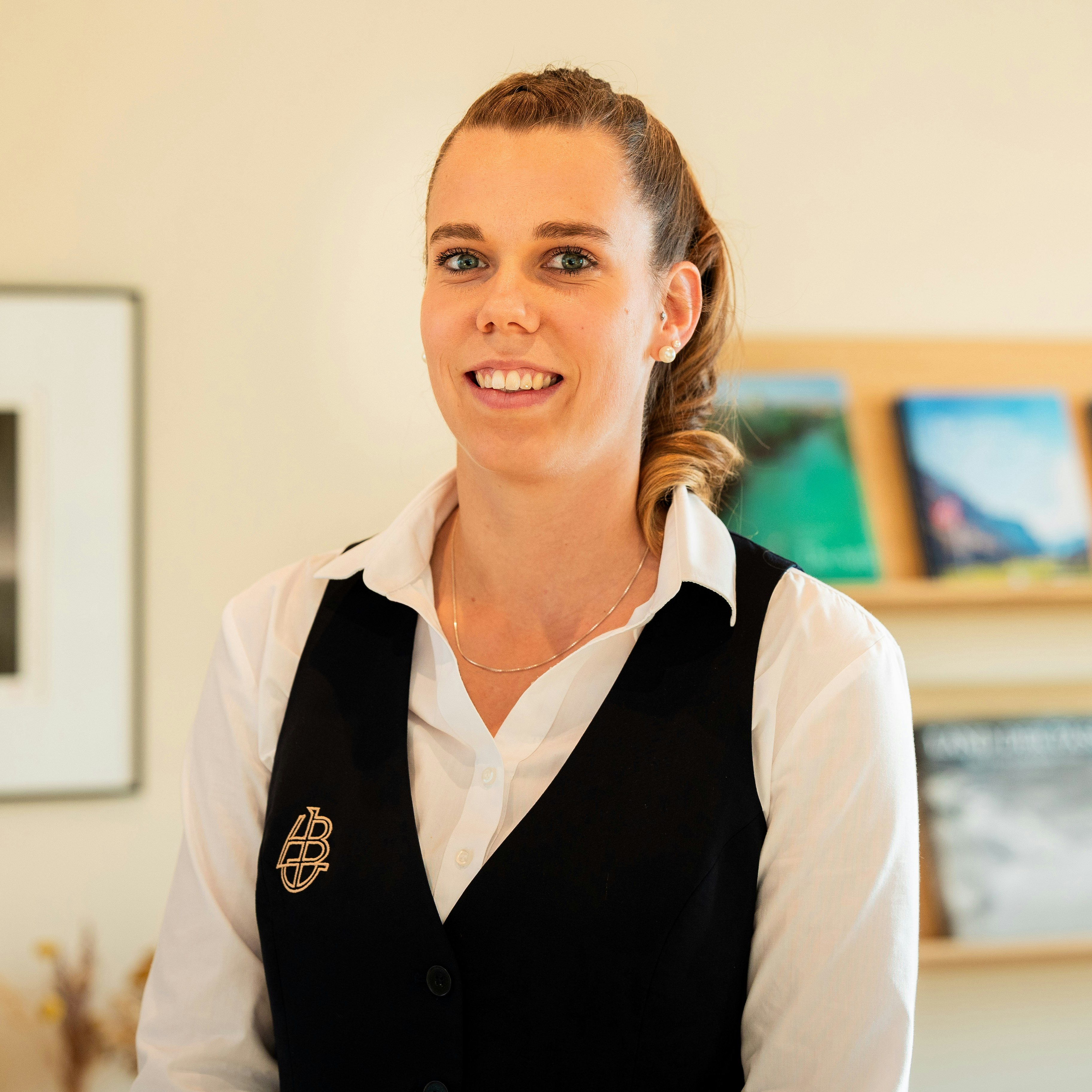 Martina Kohli, Executive Housekeeping Manager à l'hôtel BEATUS Femme souriante en chemisier blanc et gilet noir devant une étagère de magazines et une photo encadrée