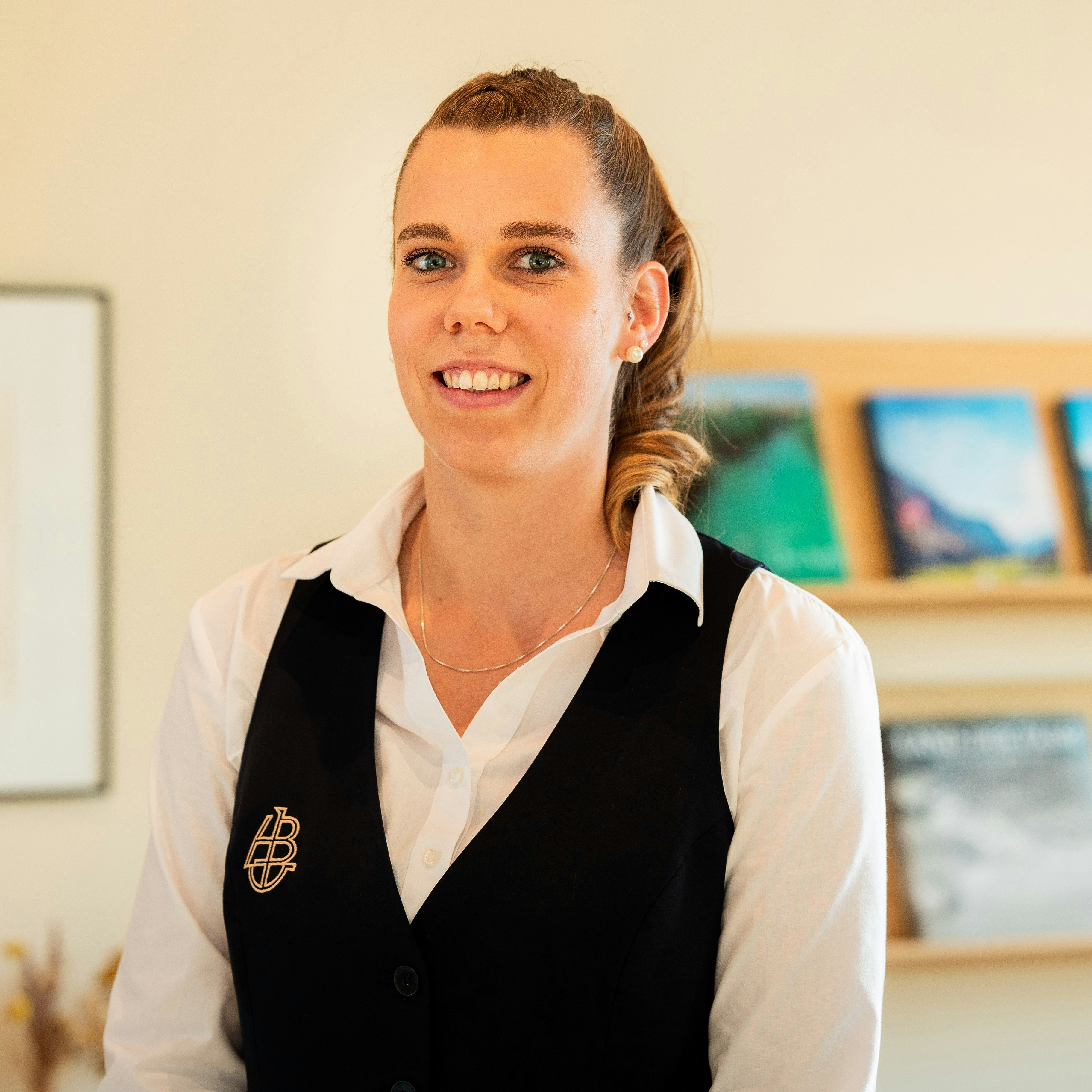 Martina Kohli, Executive Housekeeping Manager im Hotel BEATUS Lächelnde Frau mit weisser Bluse und schwarzem Gilet vor einem Regal mit Magazinen und einem gerahmten Foto