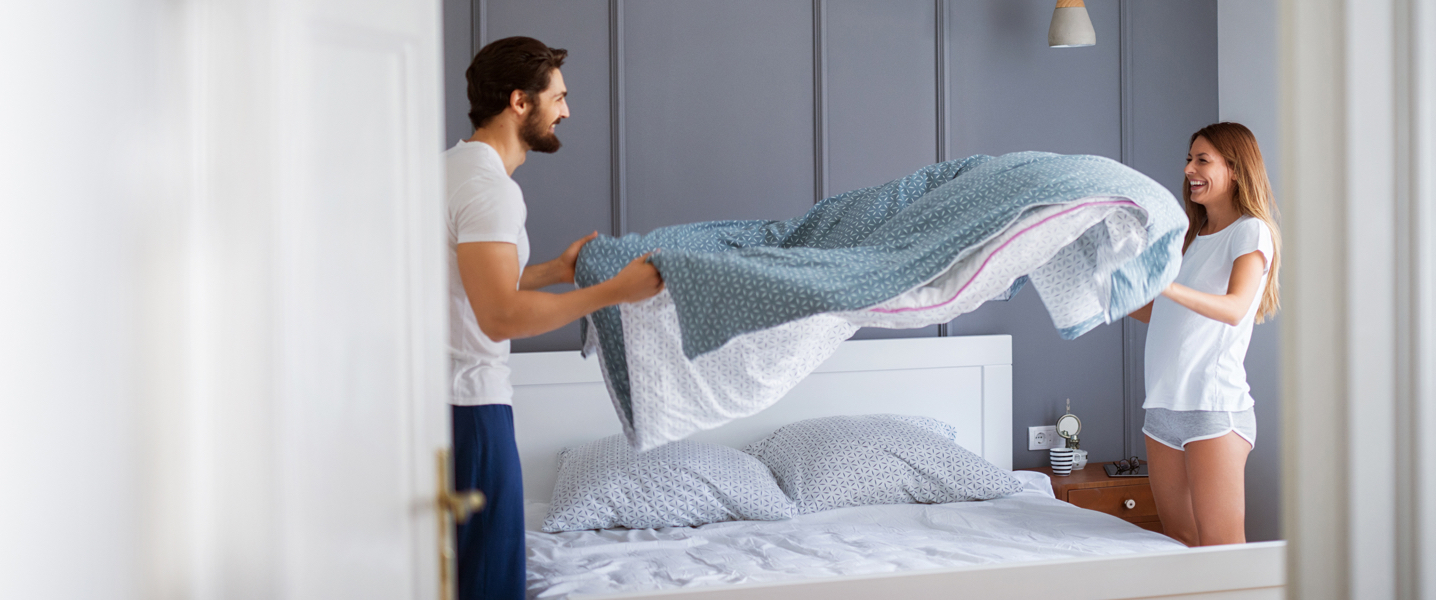 A couple smiling while they lay their blanket and sheets on their therapedic bed