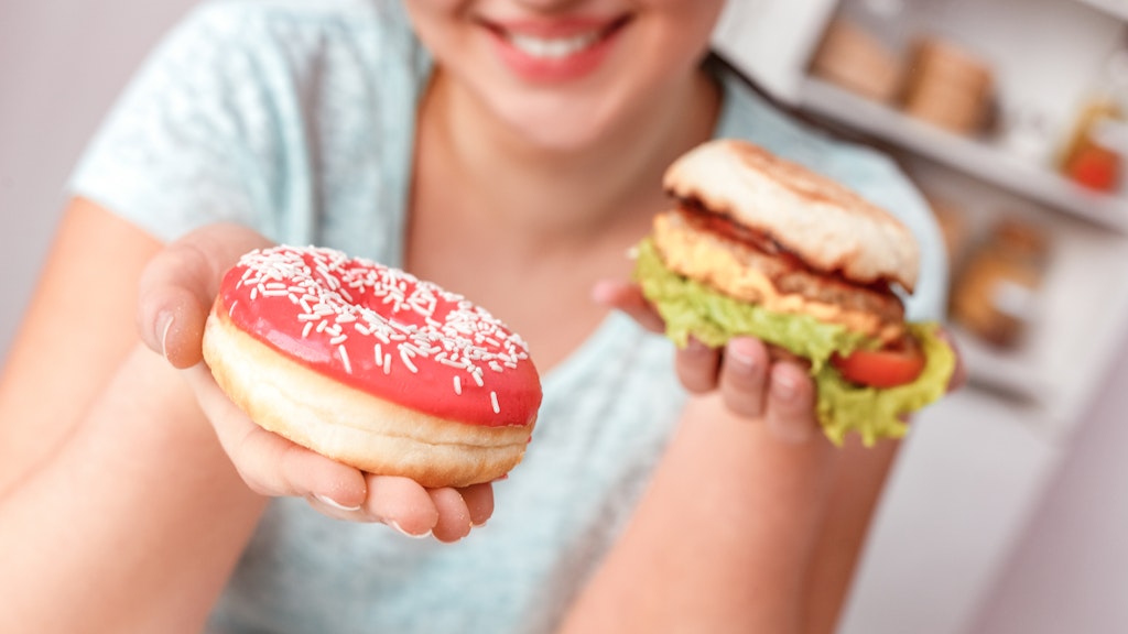 A woman with a hamburger in one hand and presenting a frosted donut with sprinkles in the other
