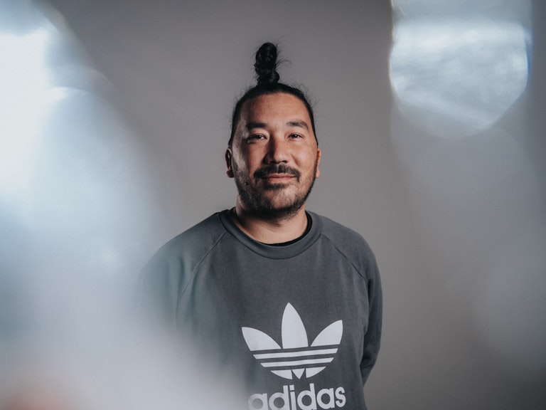 Mann mit Haarknotenfrisur in einem grauen Adidas-Sweatshirt steht vor einem neutralen Hintergrund und schaut in die Kamera.