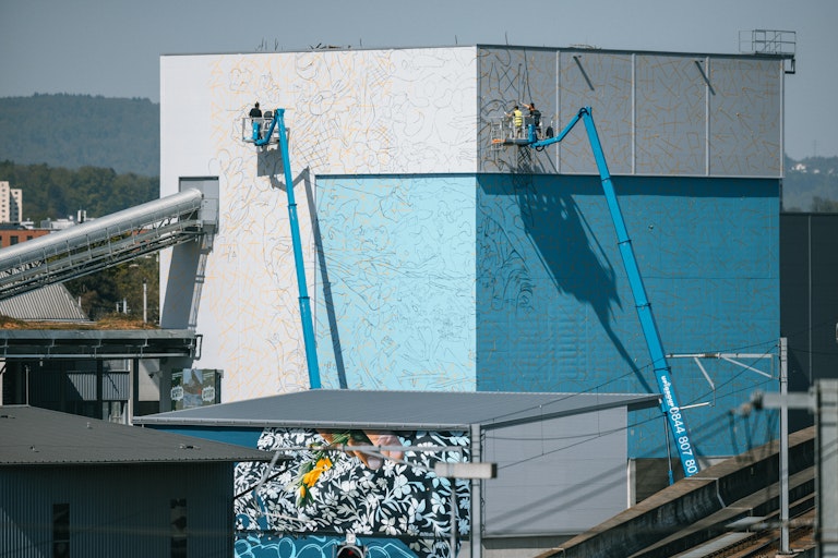Zwei Arbeiter mit Hebebühnen malen ein Wandgemälde auf ein hohes Gebäude, wobei auf den blauen und weißen Wänden umrissene Muster zu sehen sind.