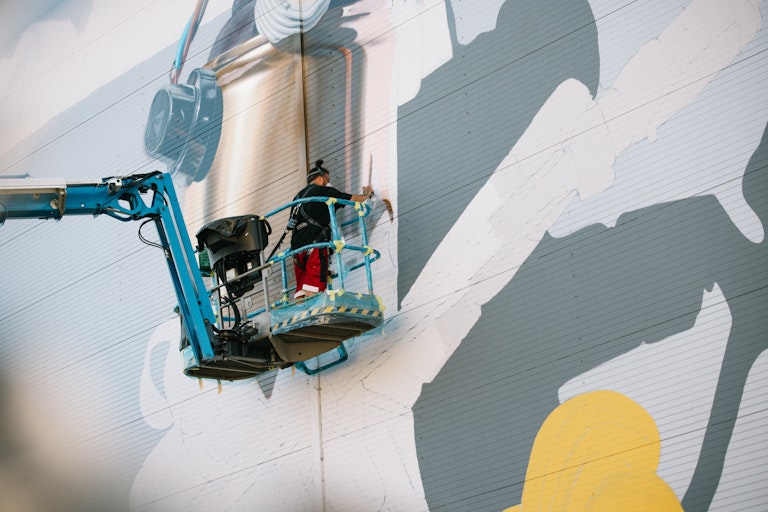 Eine Person in einem blauen Aufzug malt ein großes Wandgemälde an eine Wand und verwendet Sprühfarbe, um dem Kunstwerk feine Details hinzuzufügen.