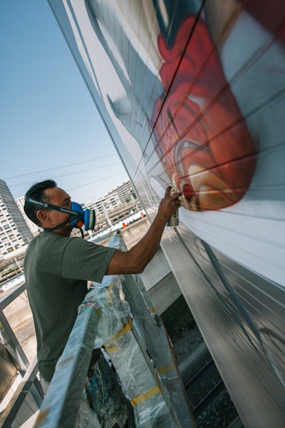 Eine Person mit Atemschutzmaske sprüht ein Wandbild auf eine große Außenwand, während sie auf einem Gerüst steht.